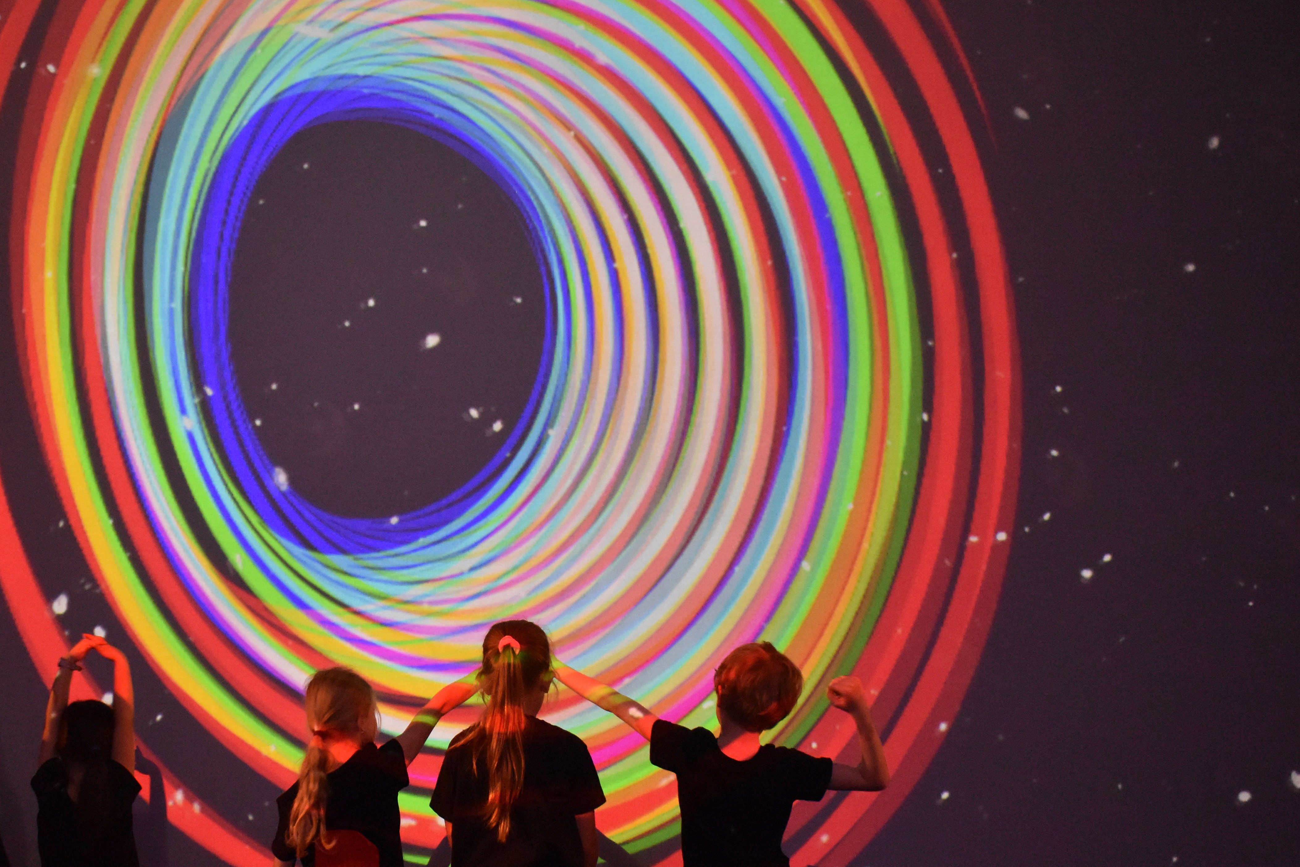 Kinder stehen vor einer bunten Lichtinstallation mit spiralförmigen Farben und einem sternenartigen Hintergrund.