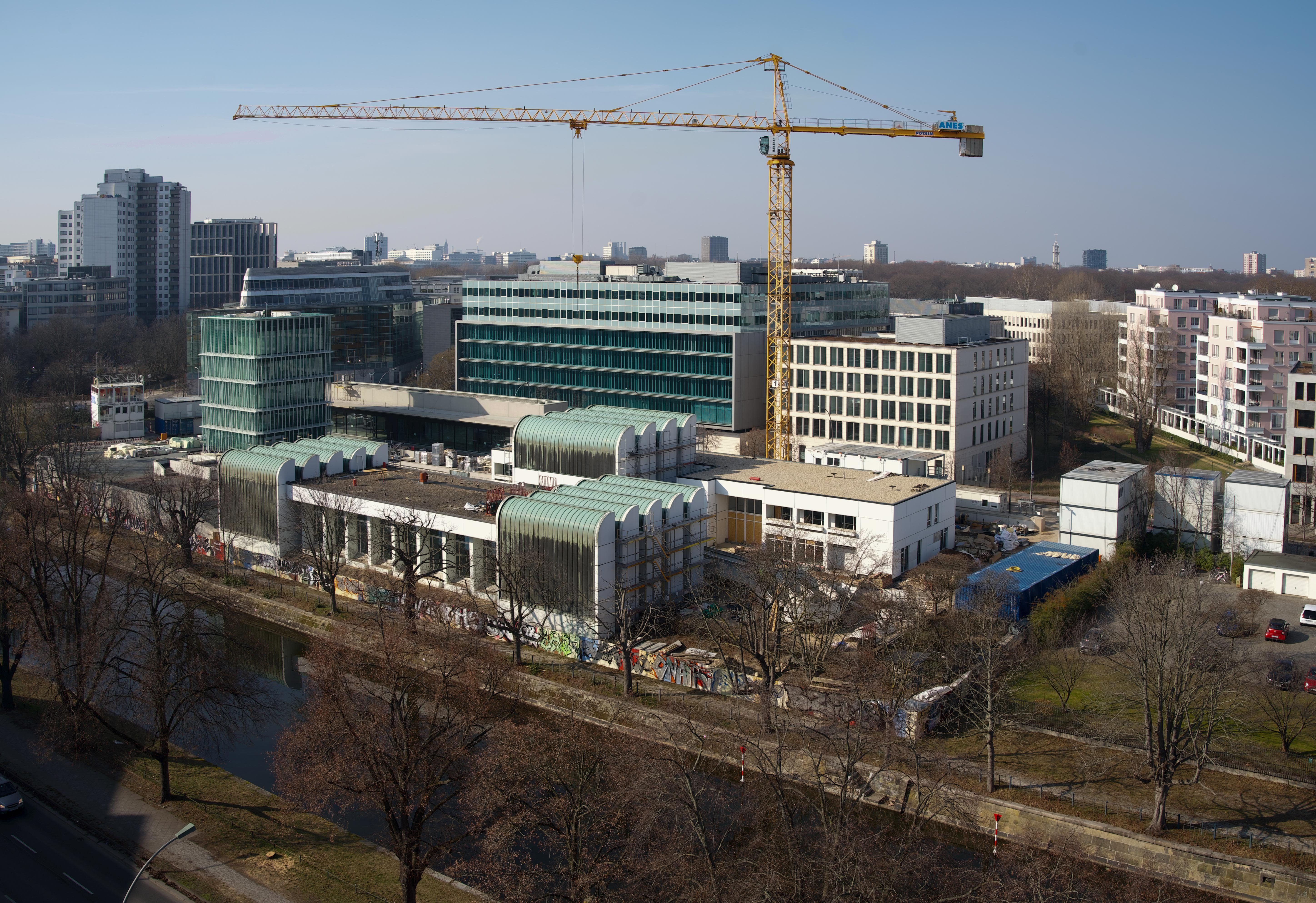 Bauprojekt mit Kränen und modernen Gebäuden, umgeben von einem Park und städtischer Umgebung an einem klaren Tag.