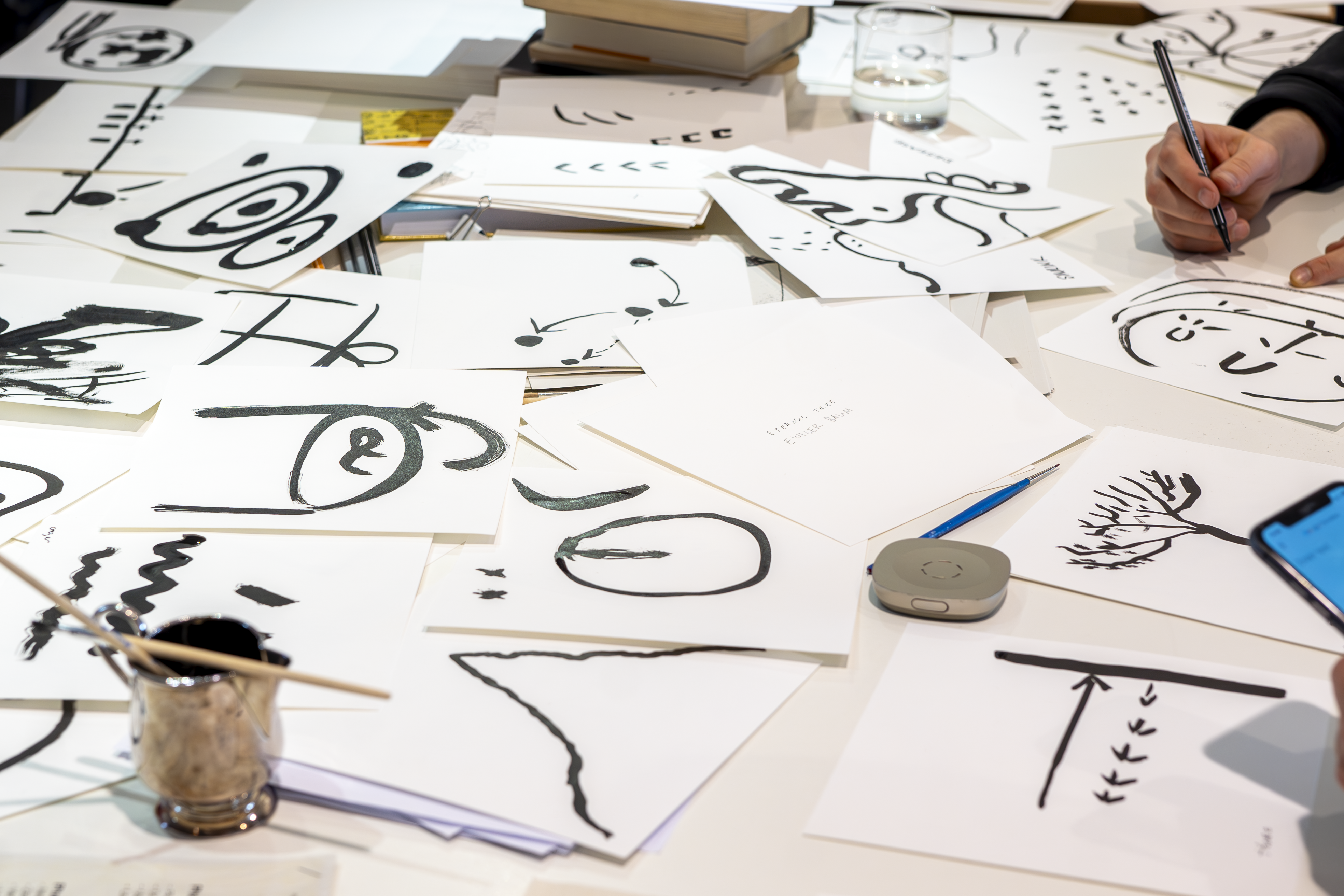 Tisch mit Zeichnungen und Skizzen in schwarzer Tinte, im Hintergrund eine Hand mit Stift und Wasserglas.