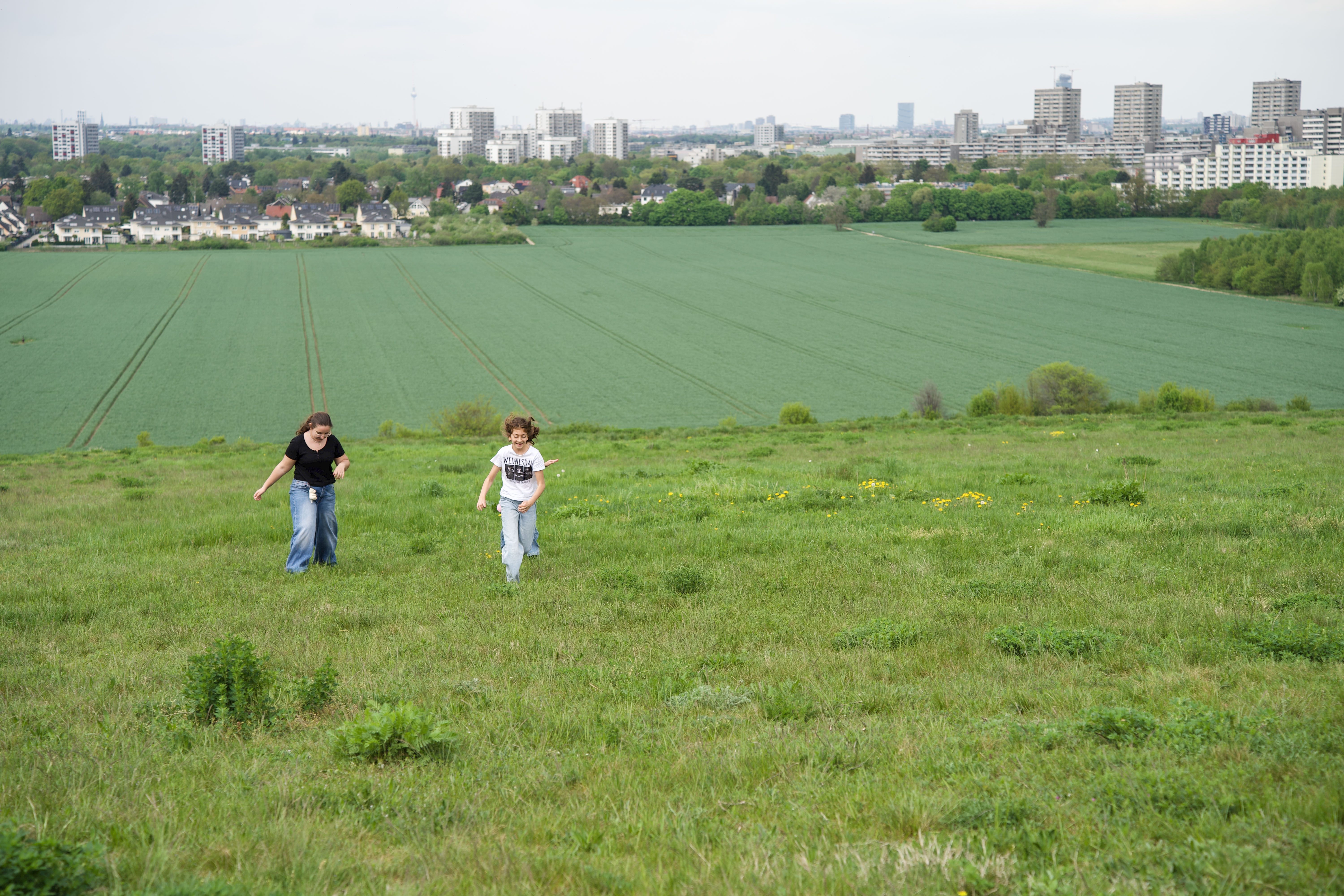 Zwei Kinder laufen fröhlich über eine grüne Wiese, im Hintergrund ist eine städtische Landschaft zu sehen.