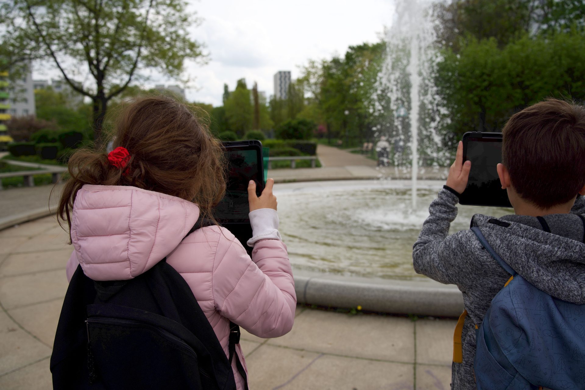 Zwei Kinder fotografieren mit Tablets einen Sprudelbrunnen im Stadtpark, im Hintergrund stehen grüne Bäume.