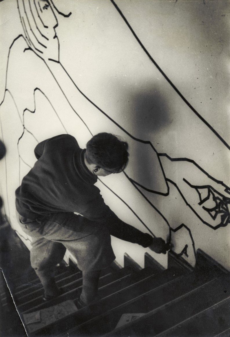 An artist paints a wall with abstract lines while standing on a ladder. Black-and-white photo.