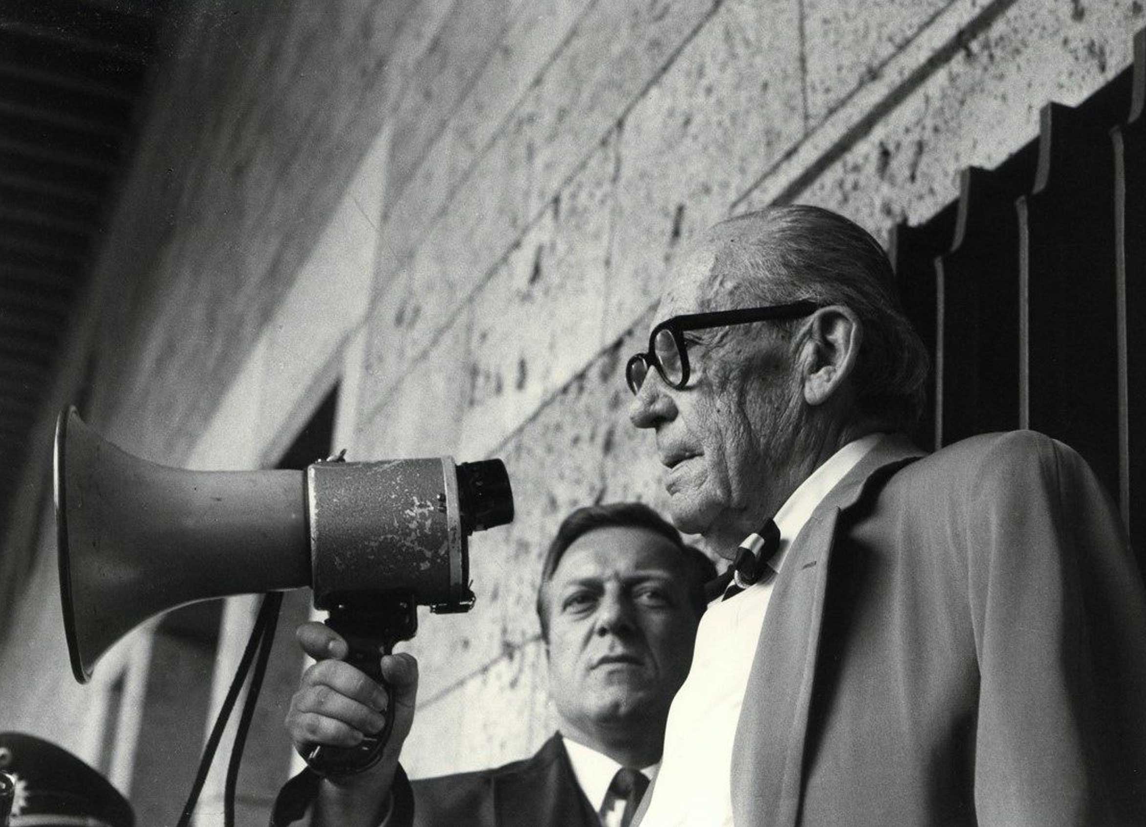 Two men stand at a microphone, one speaking while the other listens attentively. Black and white photo.