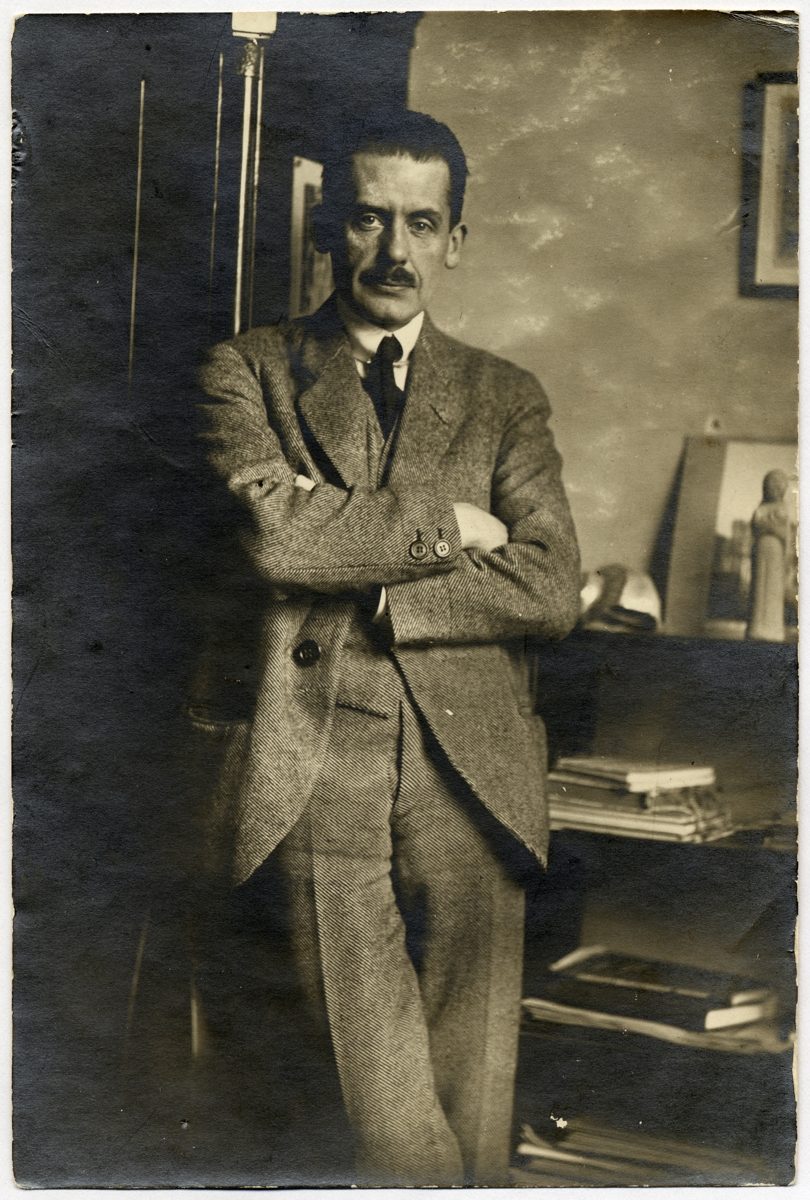 Black-and-white portrait of a man in a suit, standing with crossed arms in a room filled with books and pictures.