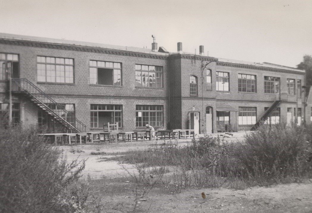 Black-and-white image of a faded brick building with windows and steps, featuring outdoor tables in front.