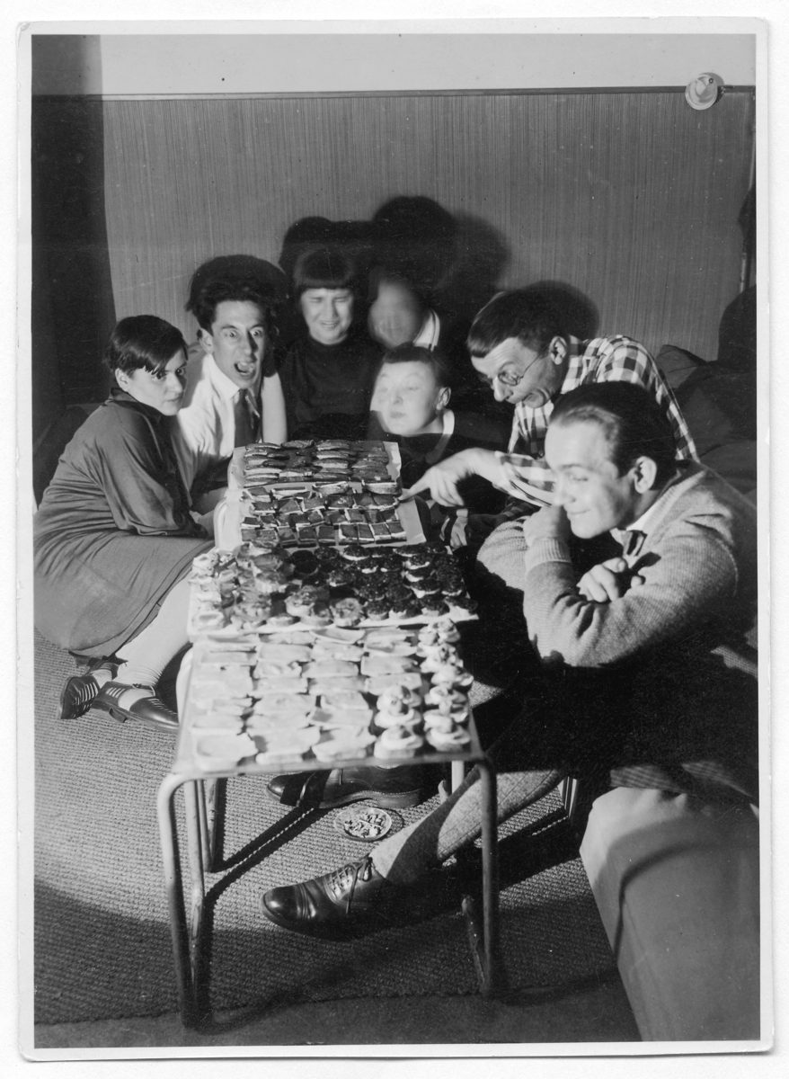 A group of people sits around a table with various treats in a cozy room. Black and white photo.