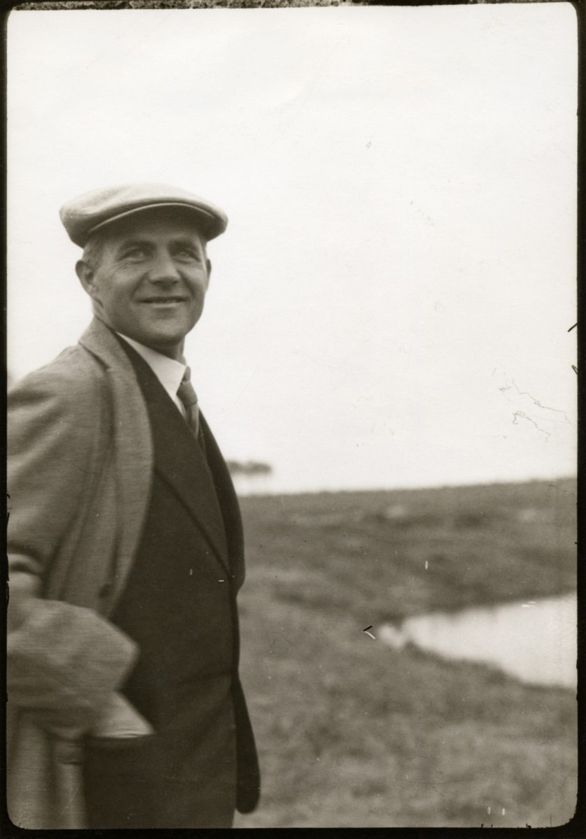 A man in a hat and suit smiles in front of a landscape with water and grass. Nostalgic black-and-white photo.