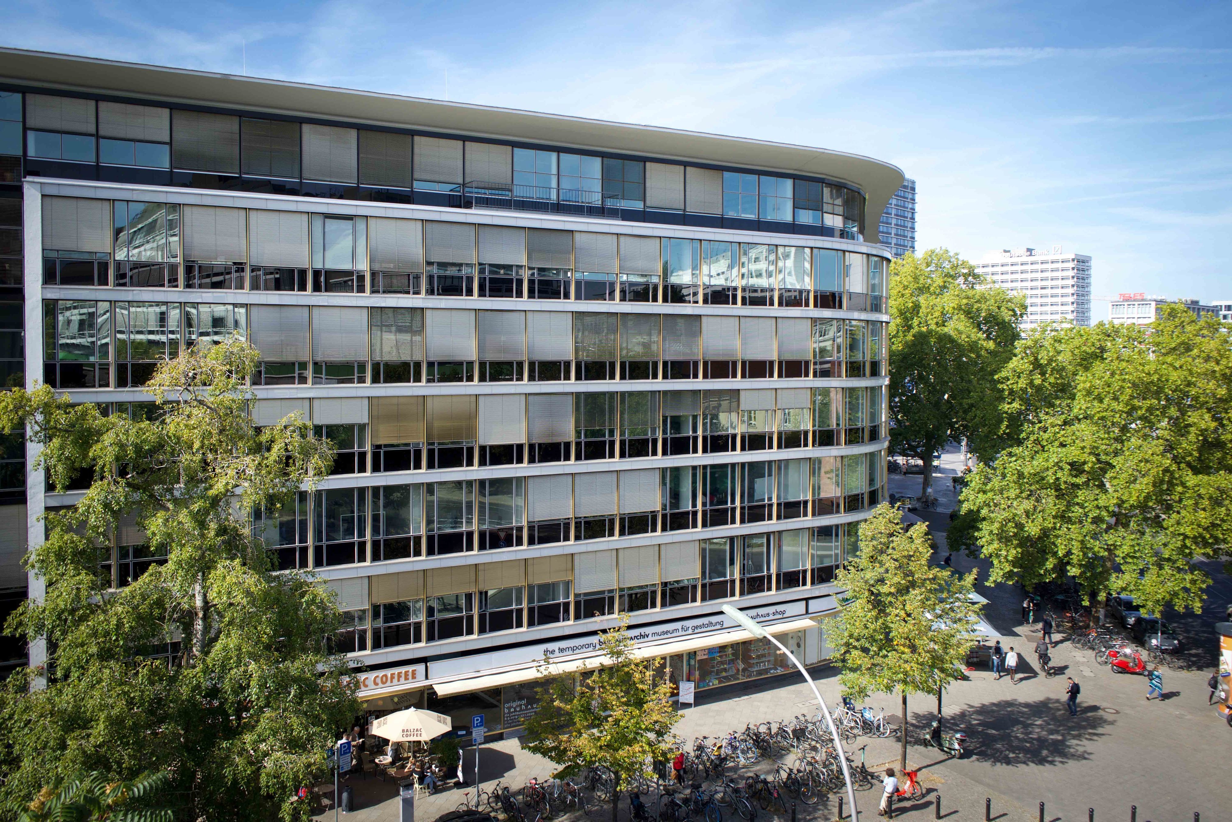 Moderne Gebäude mit großen Fenstern, Bäumen und einem Café im Erdgeschoss, belebte Straße mit Radfahrern und Fußgängern.