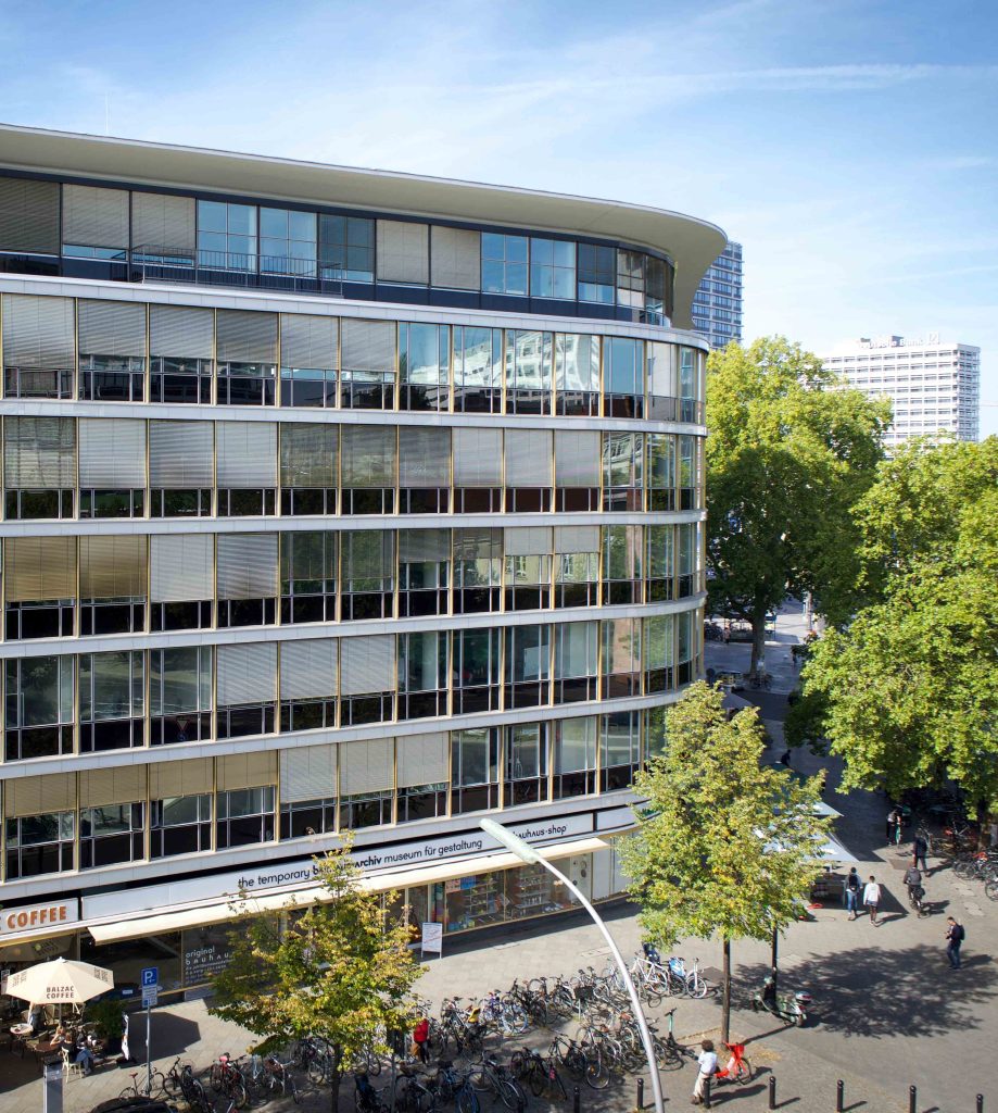 Moderne Gebäude mit großen Fenstern, Bäumen und einem Café im Erdgeschoss, belebte Straße mit Radfahrern und Fußgängern.