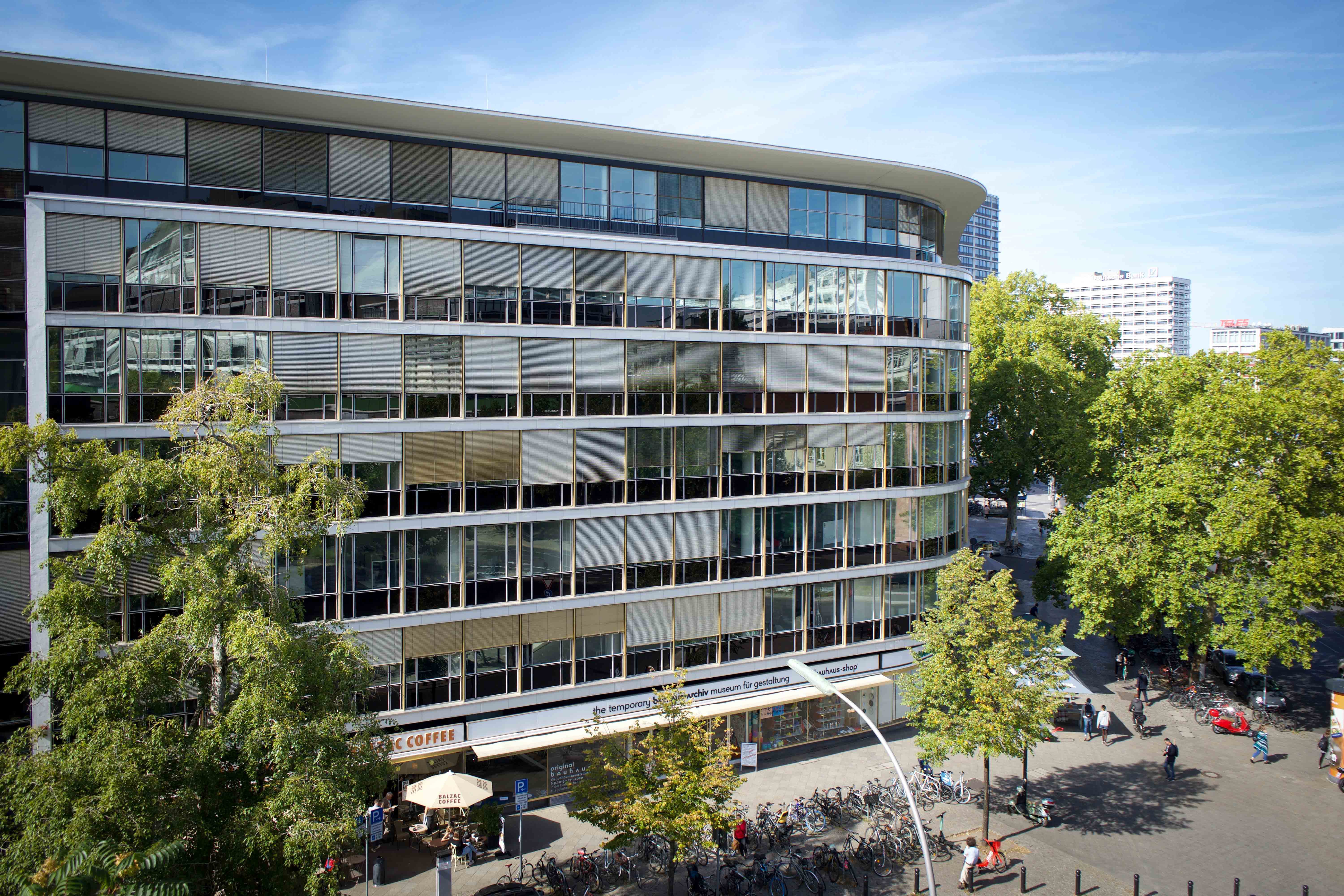 Modern building with large windows, trees, and a café on the ground floor, lively street with bicycles and pedestrians.