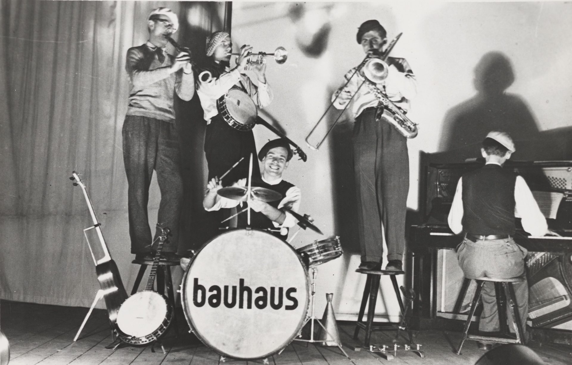 Das Foto zeigt eine fünfköpfige Musikgruppe. In der Mitte des Bildes sitzt ein Mann am Schlagzeug, am rechten Bildrand einer am Klavier. Auf der Schlagzeugtrommel steht "bauhaus". Drei Musiker stehen auf Stühlen. Der Erste von links spielt Klarinette, der Zweite Trompete und der Dritte Posaune.