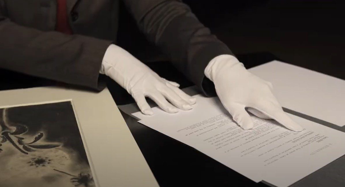 Eine Hand mit weißen Handschuhen zeigt auf ein Dokument, während sie neben einem Kunstwerk auf einem Tisch liegt.
