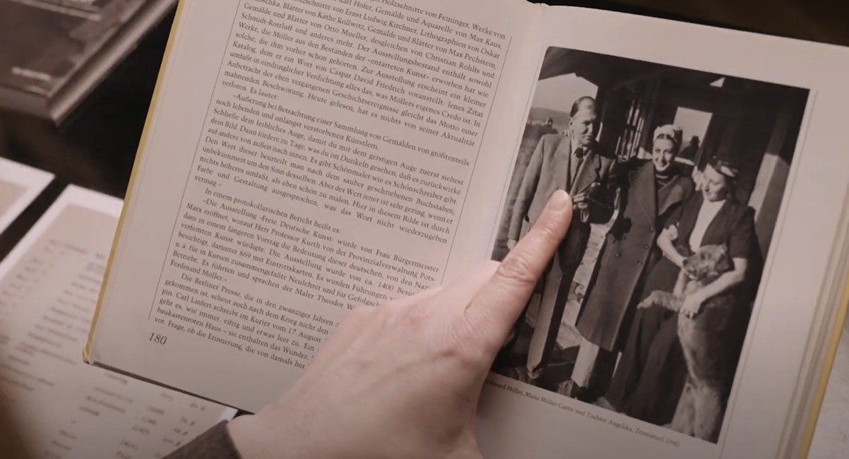 Eine Hand zeigt auf ein Foto in einem Buch, das drei Personen und einen Hund darstellt, daneben steht ein Text über Kunst.