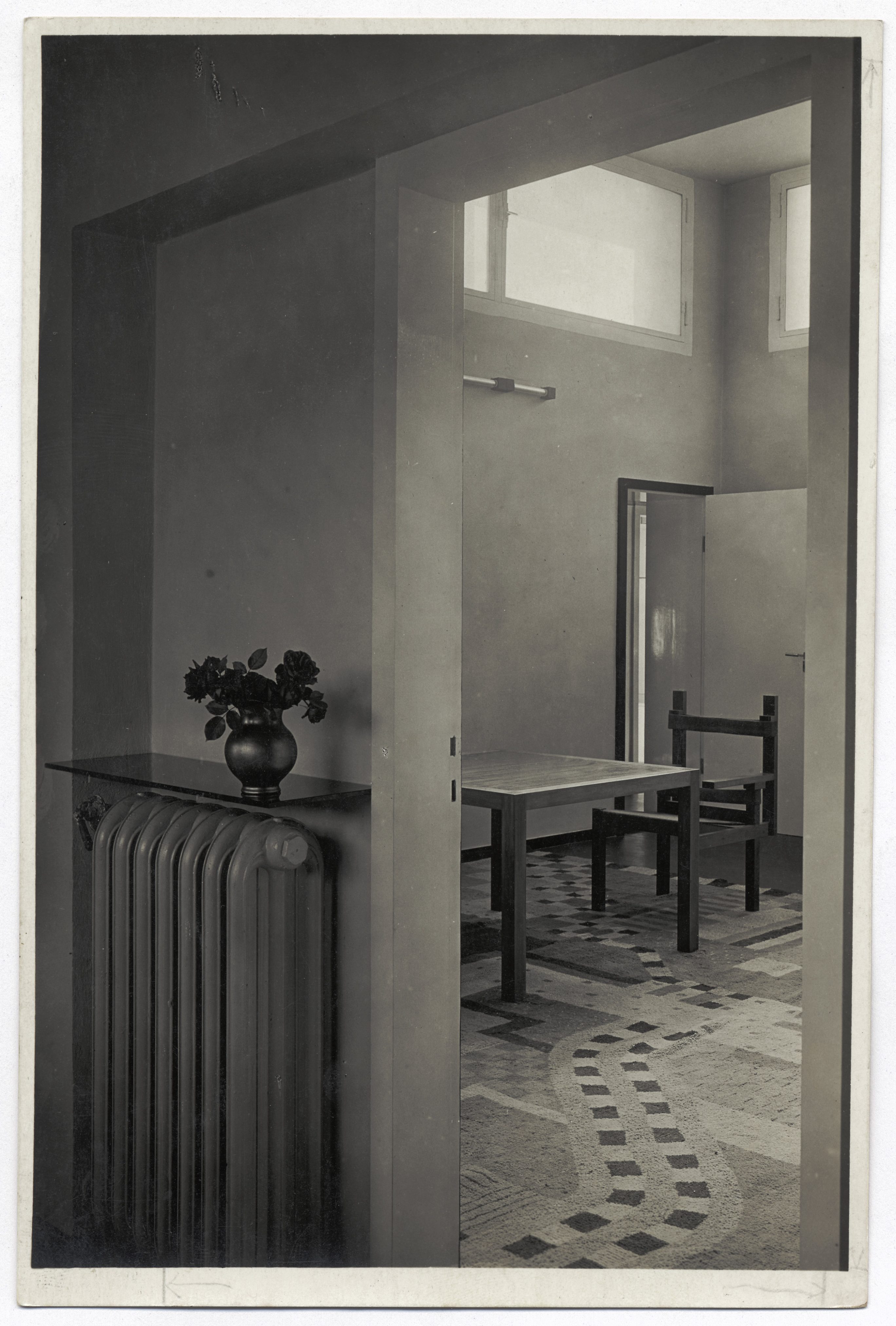 Ein minimalistischer Raum mit Tisch, Stuhl und Vase auf einem Heizkörper, sanft beleuchtet.