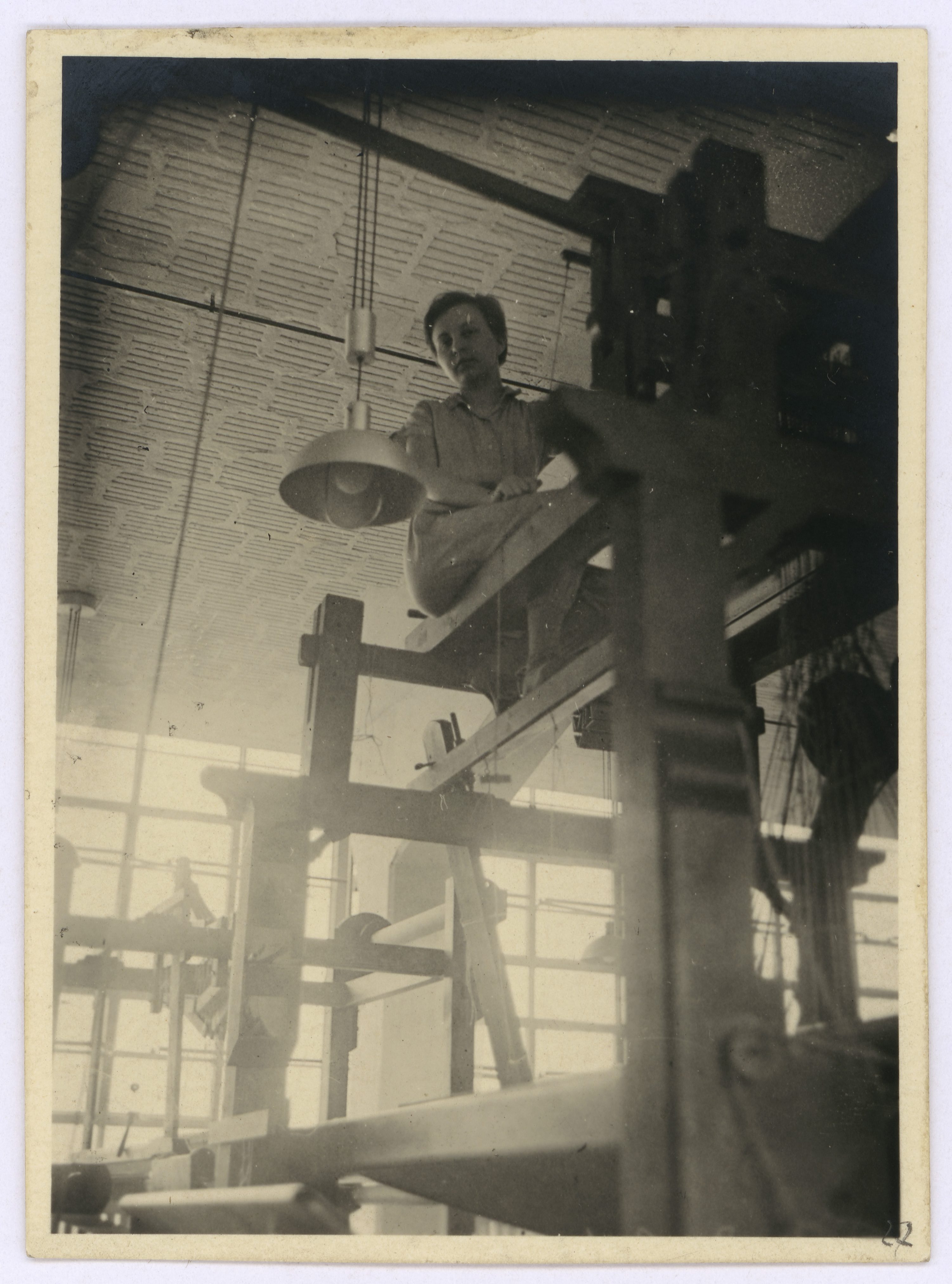 Ein Mann sitzt auf einer Holzstruktur in einer Werkstatt mit großen Fenstern und einer Lampe über ihm.