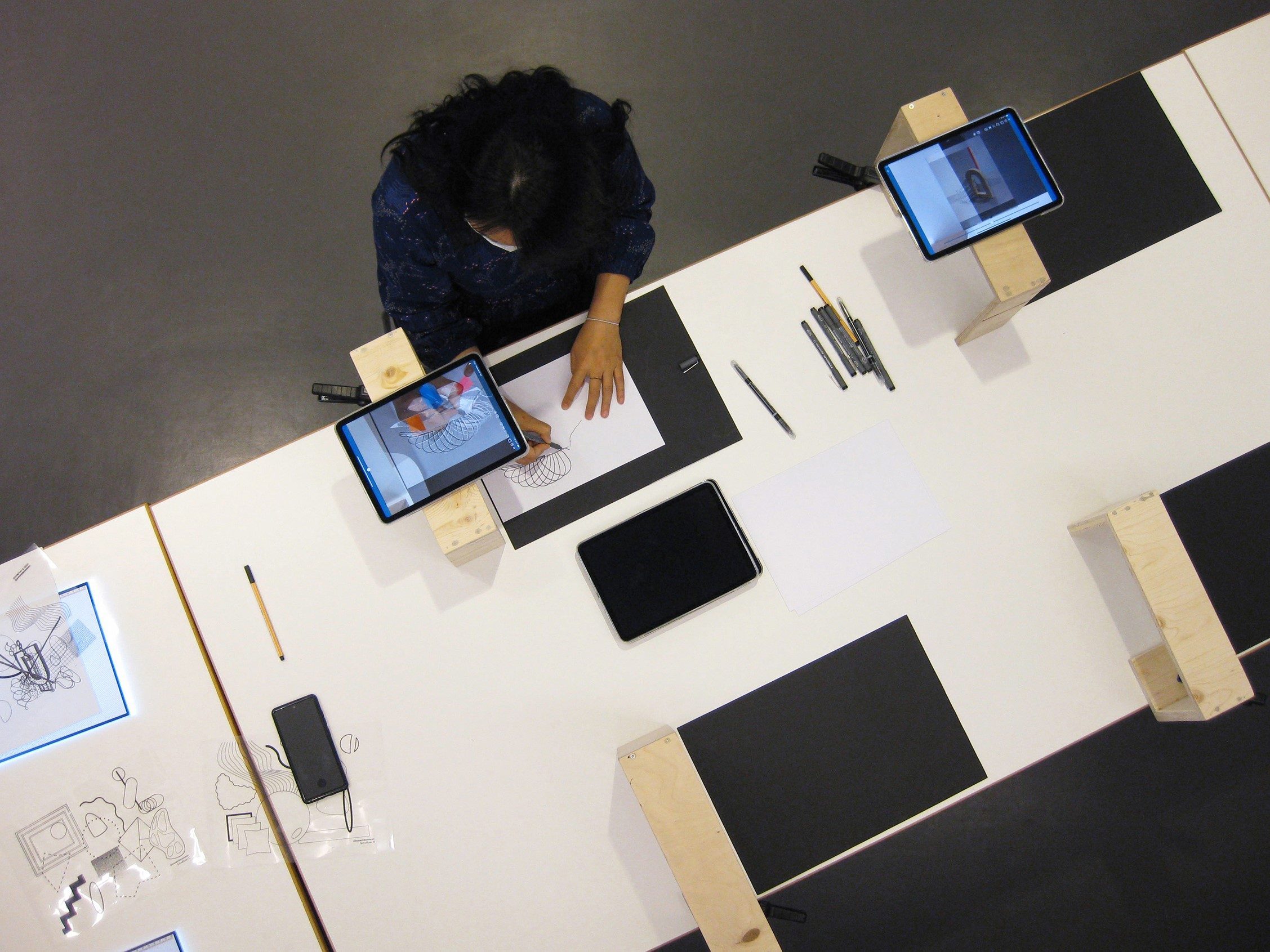 Eine Person zeichnet an einem Tisch, umgeben von Tablets, Stiften und Zeichenmaterial.