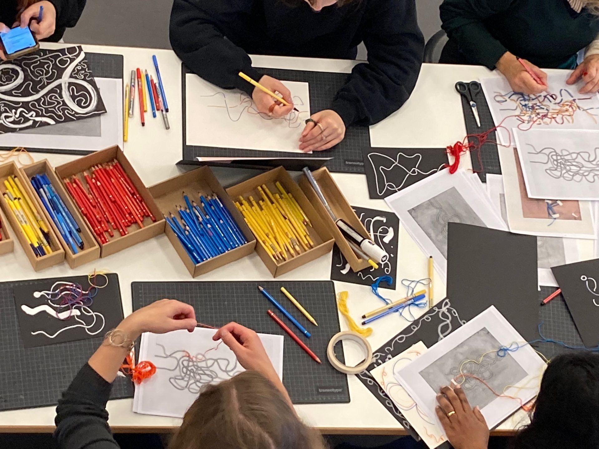 A table with creative materials, drawings, and several people working on art projects.