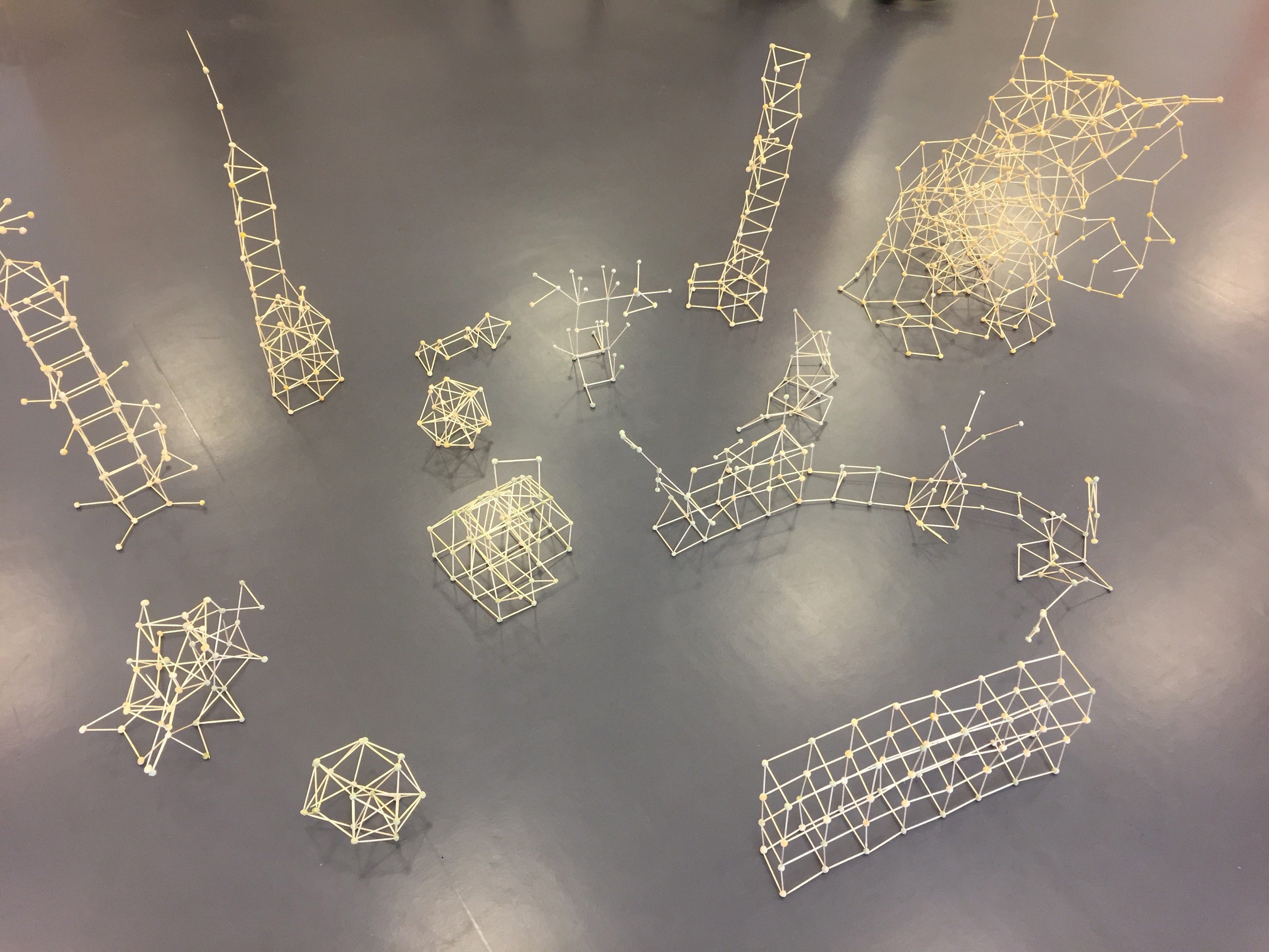 Strukturierte geometrische Modelle aus Holzstäbchen und kleinen Knoten auf einem grauen Boden kreiert.