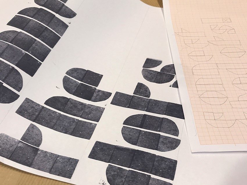 Gestapelte Papierblätter mit schwarzen Buchstabenmustern und einem skizzierten Schriftentwurf auf Karopapier.