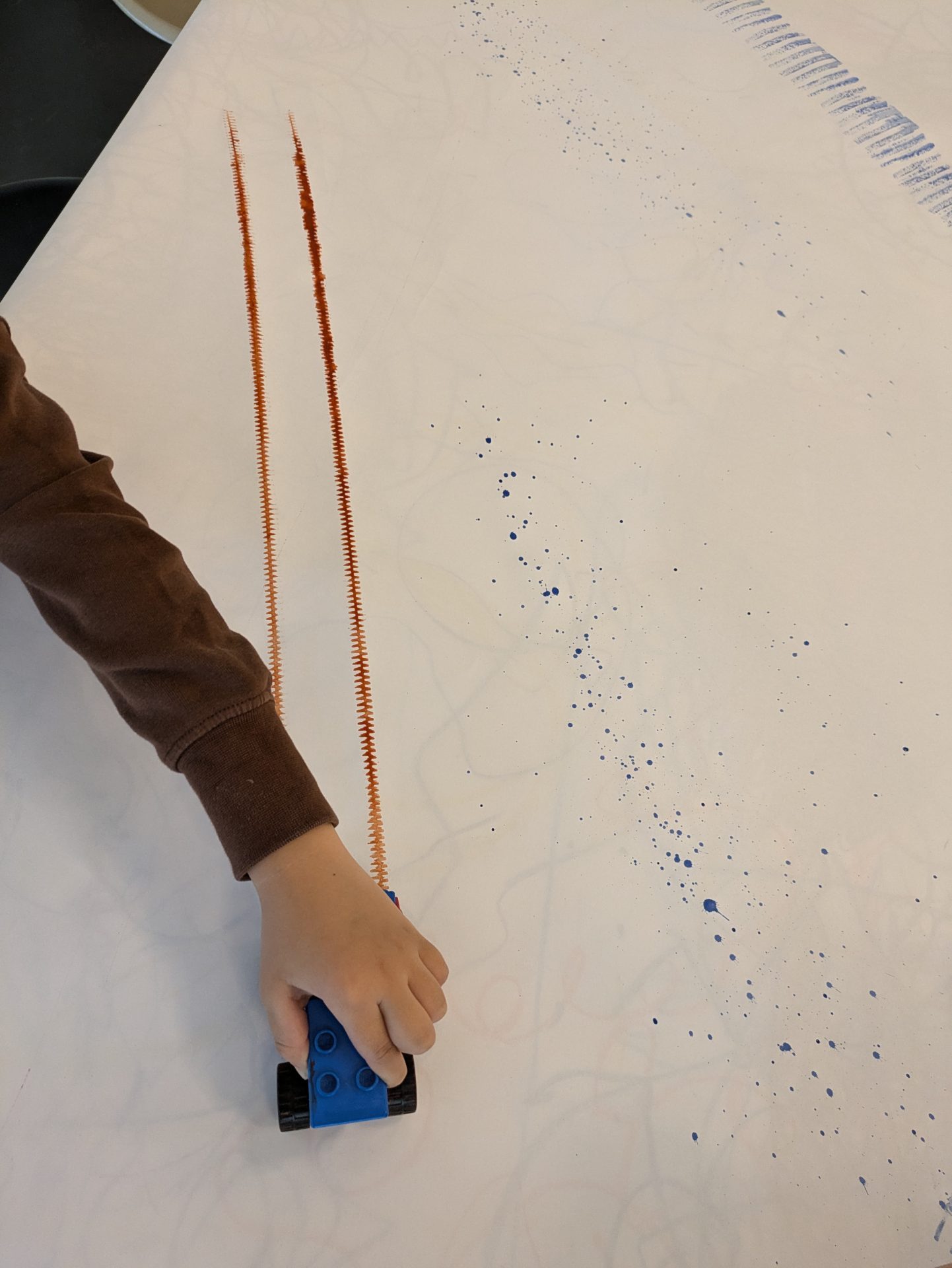 Ein Kind fährt mit einem Spielzeugauto über ein großes Blatt Papier, das mit orangefarbenen und blauen Farbresten bedeckt ist.