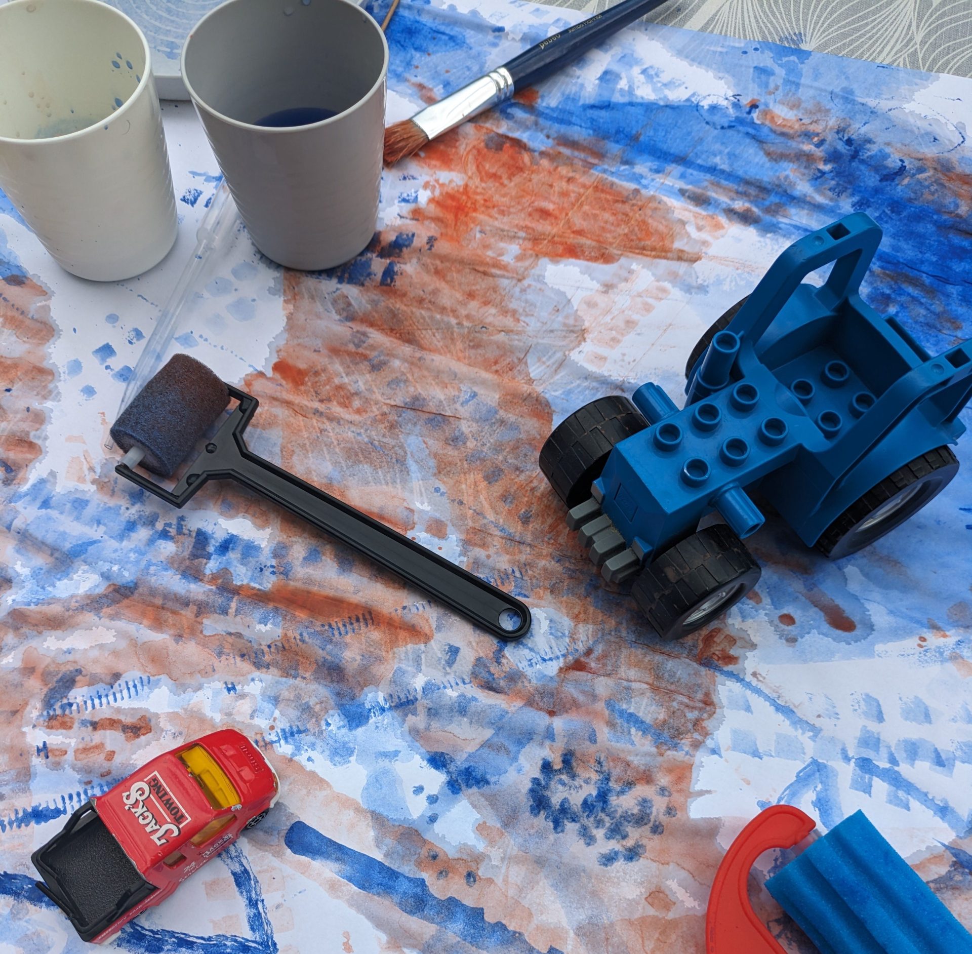 Bunte Malutensilien, darunter ein blauer Spielzeugtraktor, Farbkübel und Malwerkzeuge auf weißem Papier.
