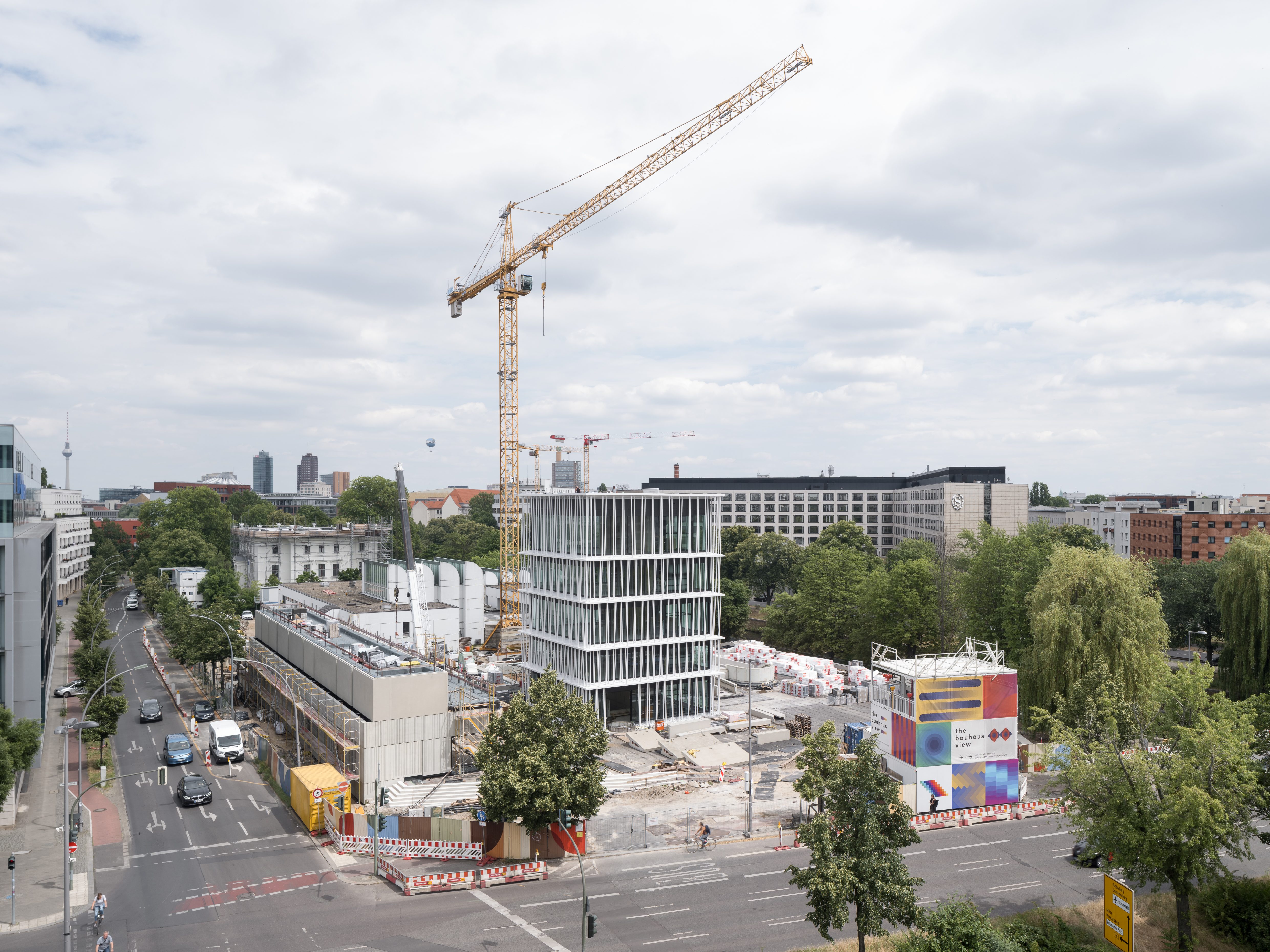 Bauarbeiten mit Kran und modernen Gebäuden in der Stadt; Wolken am Himmel, grüne Bäume.