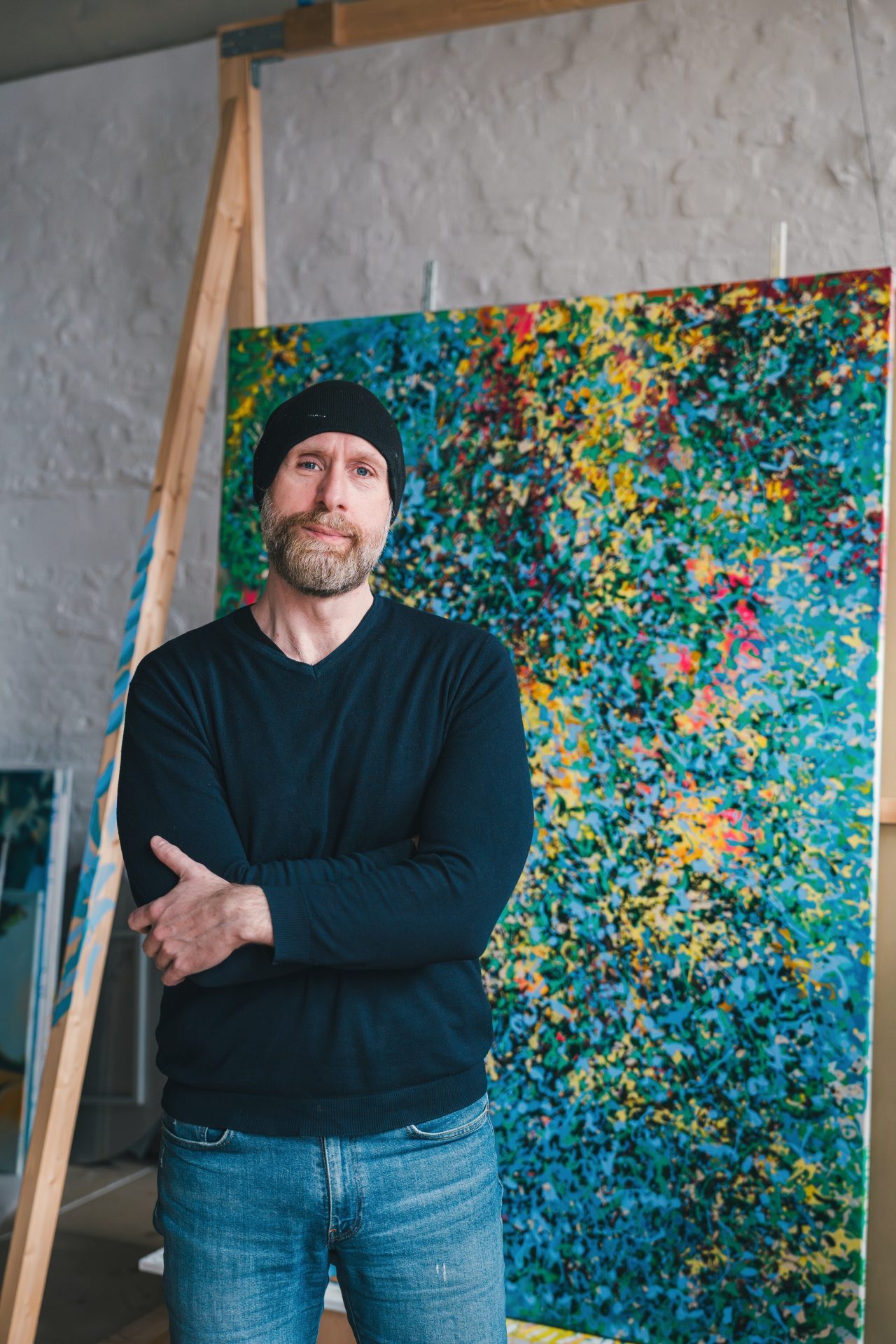 Ein bärtiger Mann mit Mütze steht vor einem bunten, abstrakten Gemälde in einem Atelier.