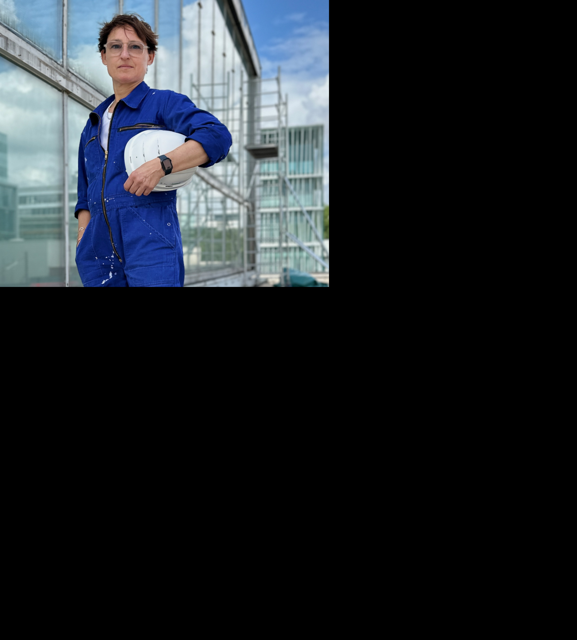 Person in blue work clothing with a helmet stands on a construction site with glass surfaces in the background.