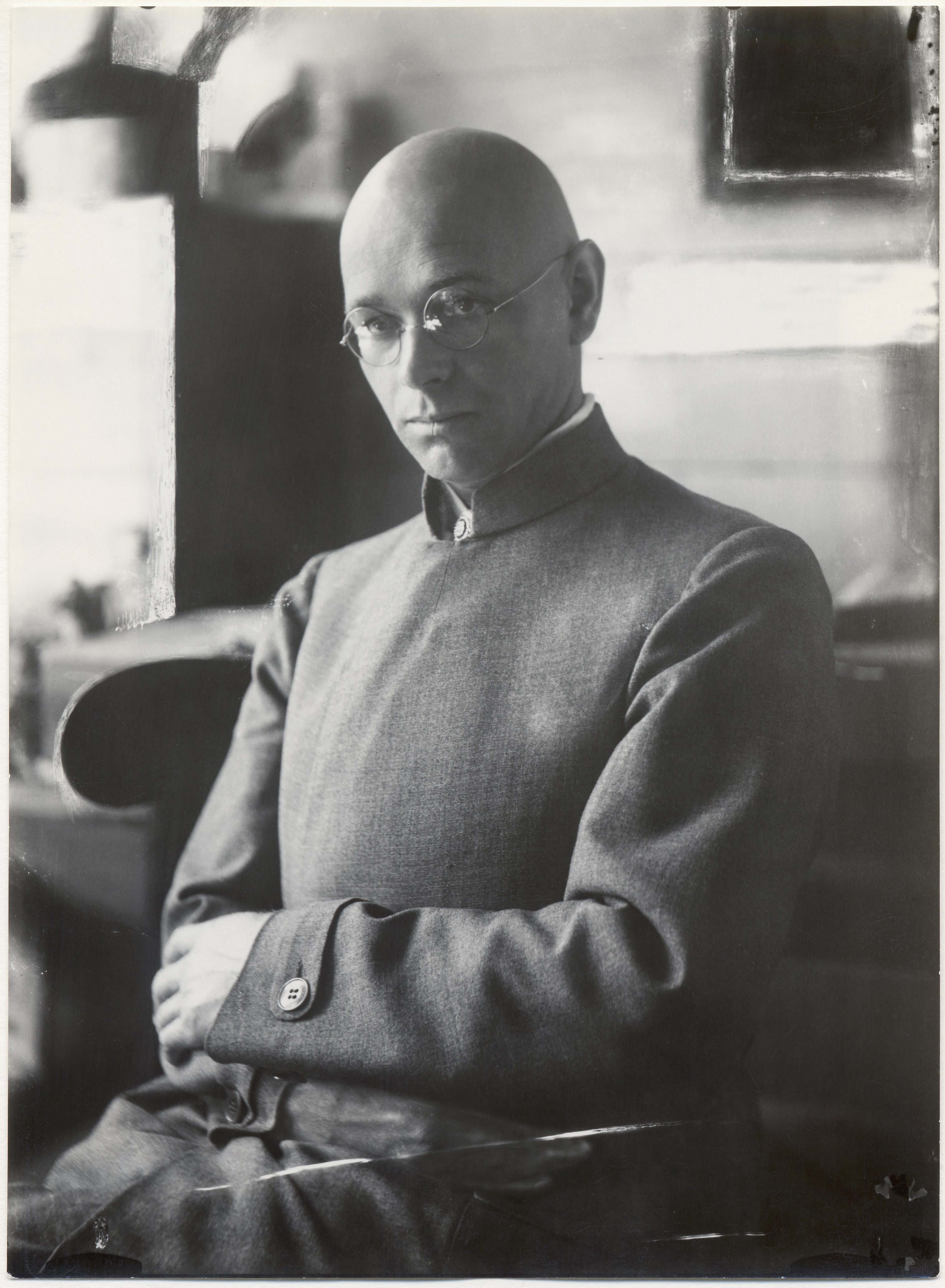 A thoughtful man in glasses and a gray suit sits with crossed arms. The background is blurred.