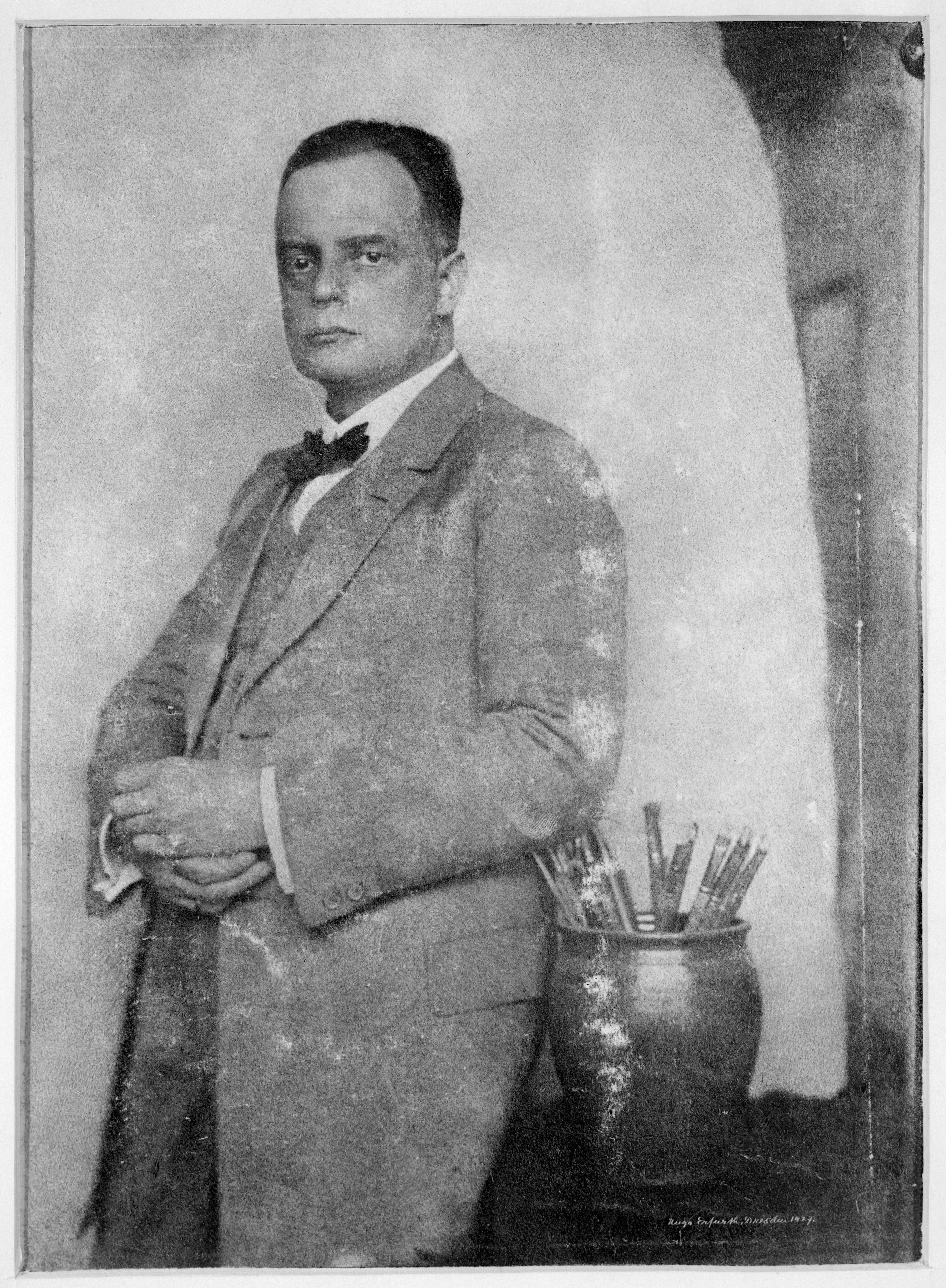 Historical portrait of a man in a suit with a bow tie, in front of a container with writing utensils.