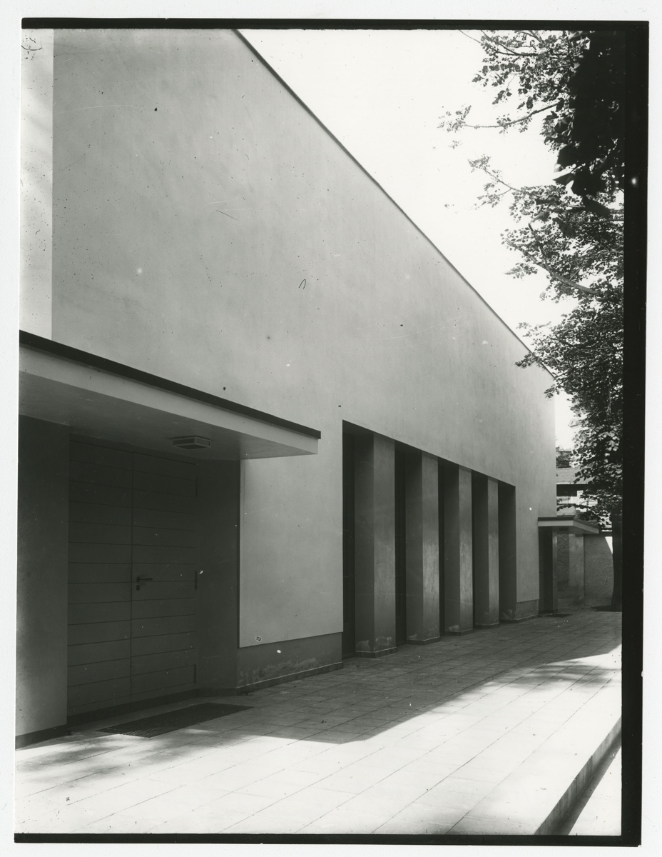 Moderner Bau mit glatter Fassade und vertikalen Elementen, im Schatten von Bäumen auf einem gepflasterten Boden.