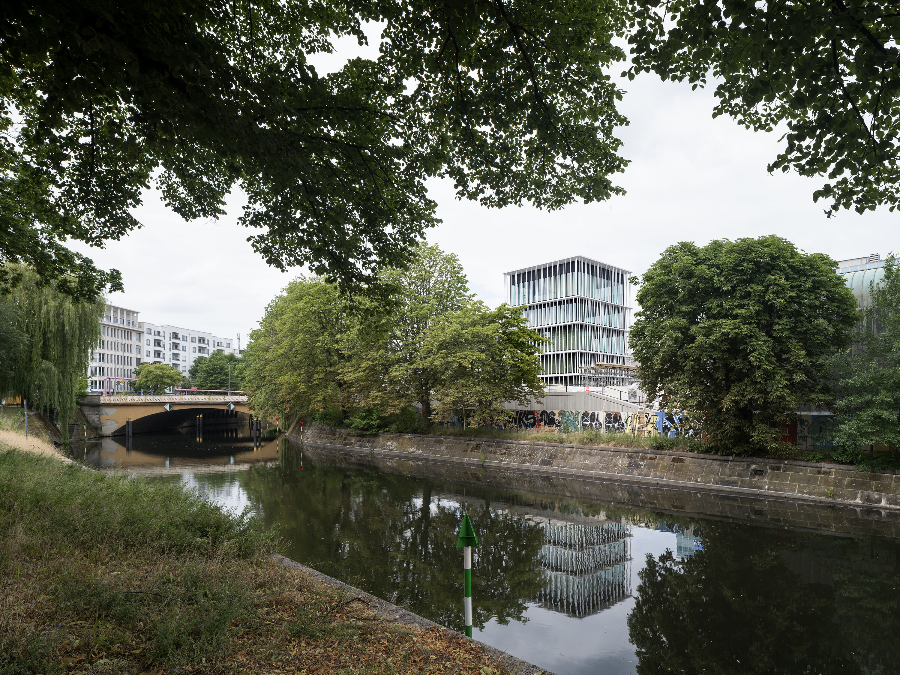 Blick auf einen Fluss mit Bäumen, einem Brückenbogen und modernen Gebäuden im Hintergrund.