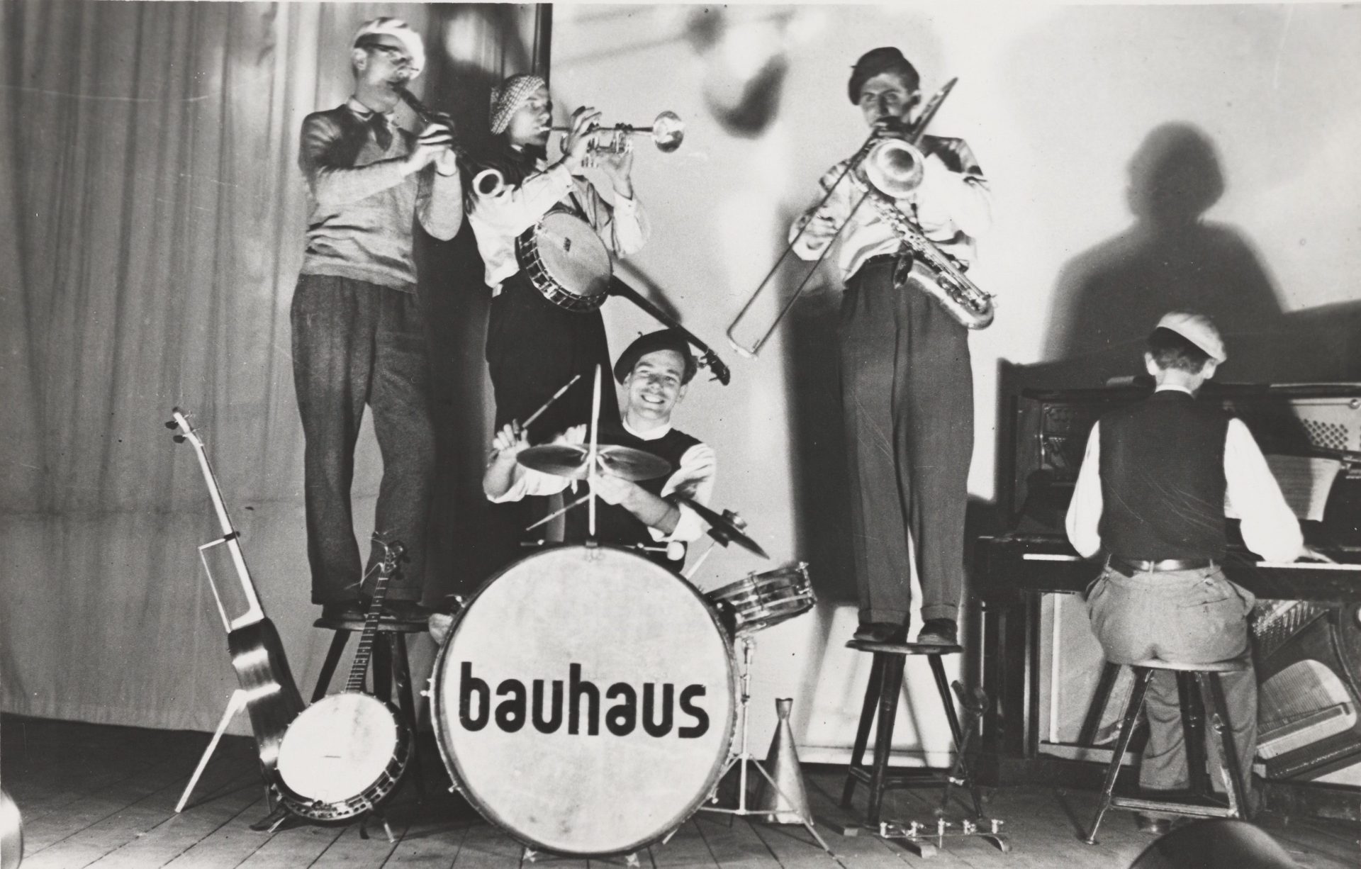 Vier Musiker in einem schwarz-weißen Bild spielen auf einer Bühne mit dem Schriftzug "bauhaus" auf dem Schlagzeug.