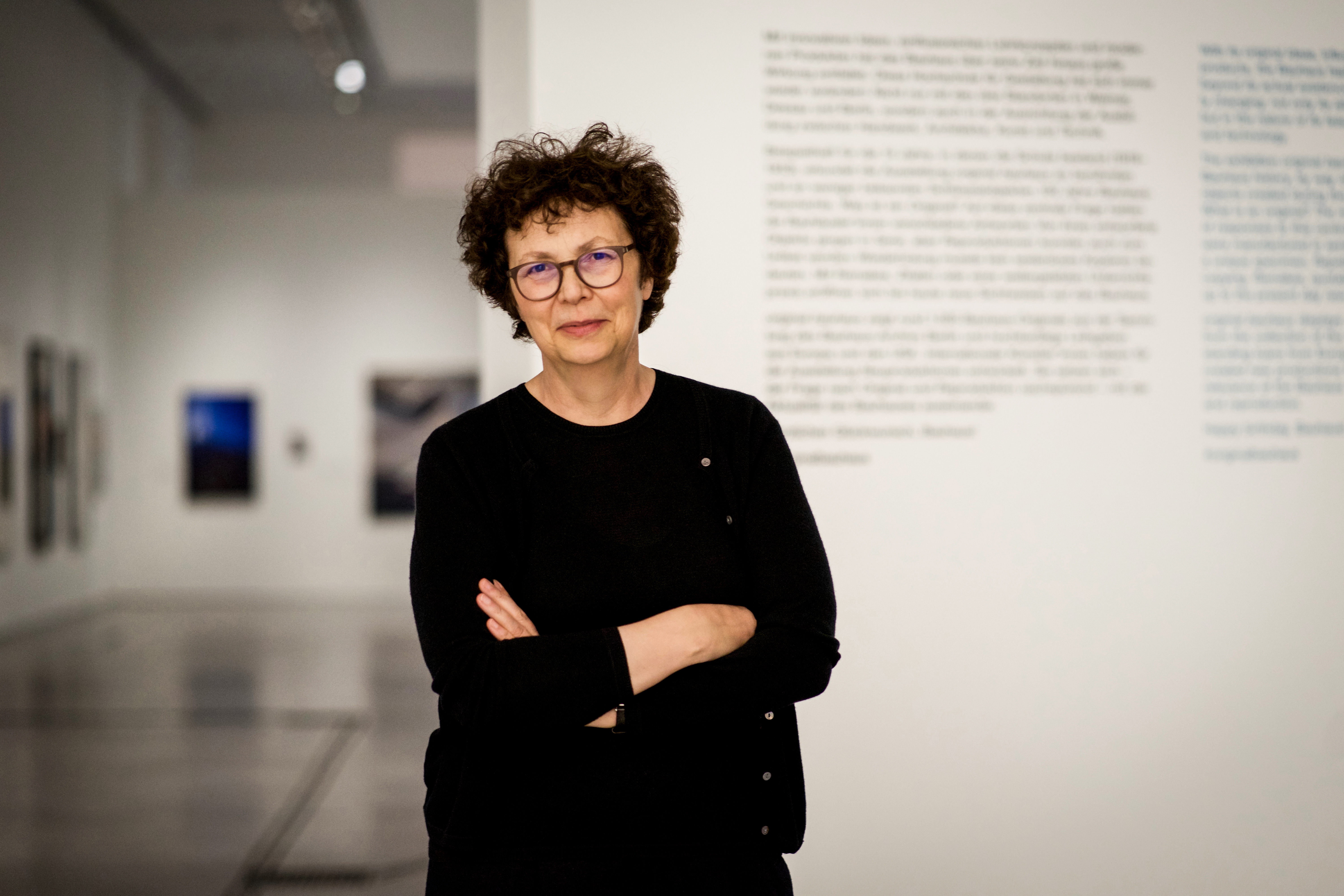 Frau mit lockigem Haar und Brille steht in einer Galerie vor einer Wand mit Schriftzügen.