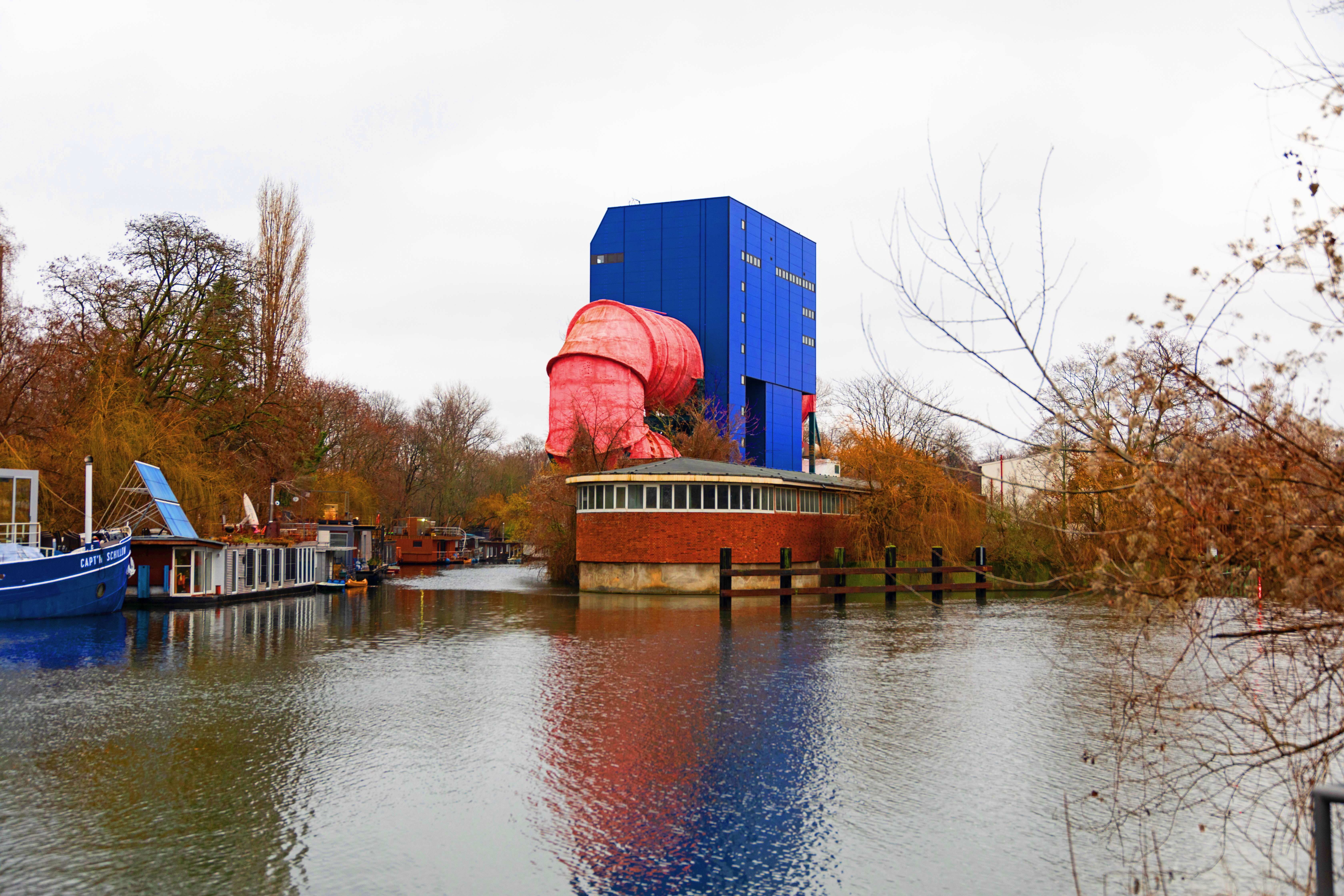 Moderne Architektur mit blauem und rotem Gebäude am Wasser, umgeben von herbstlichen Bäumen.