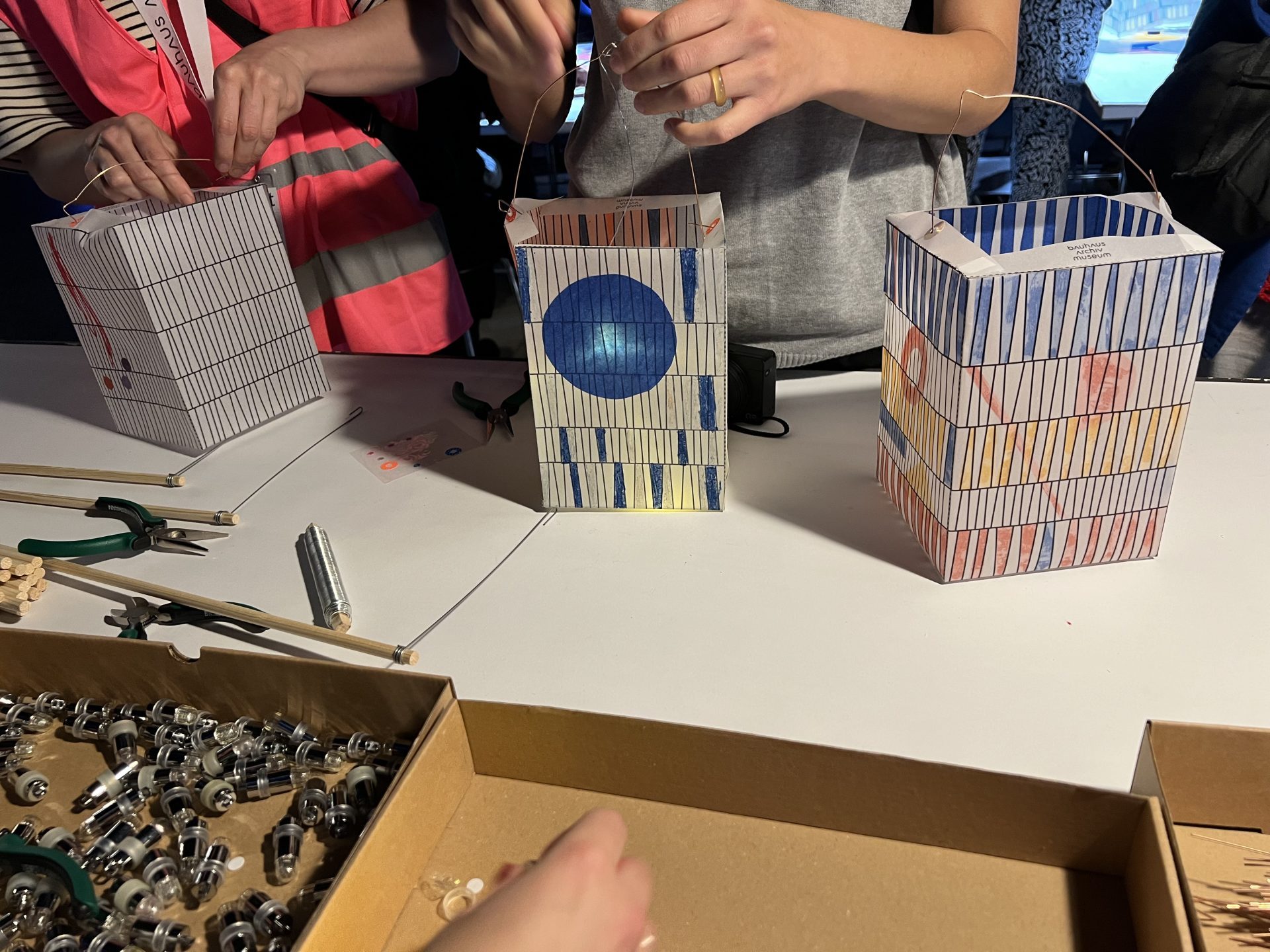 Zu sehen sind zwei bunte Laternen und einige Hände die gerade noch die letzten Schritte zum Beenden der Montage der gestalteten Laternen vornehmen. Eine Schachtel mit Befestigungsklammern ist ebenfalls zu sehen.