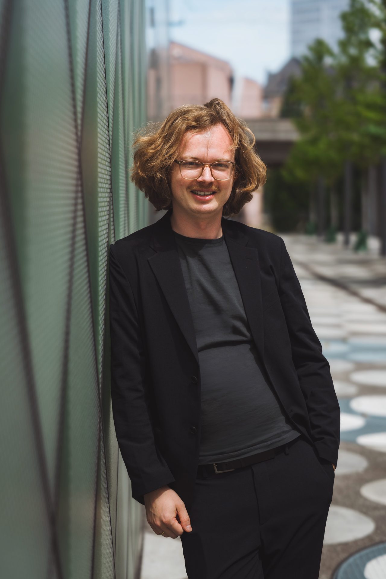 Ein Mann mit lockigem, blondem Haar und Brille in einem schwarzen Anzug lehnt entspannt an einer Wand im Freien.