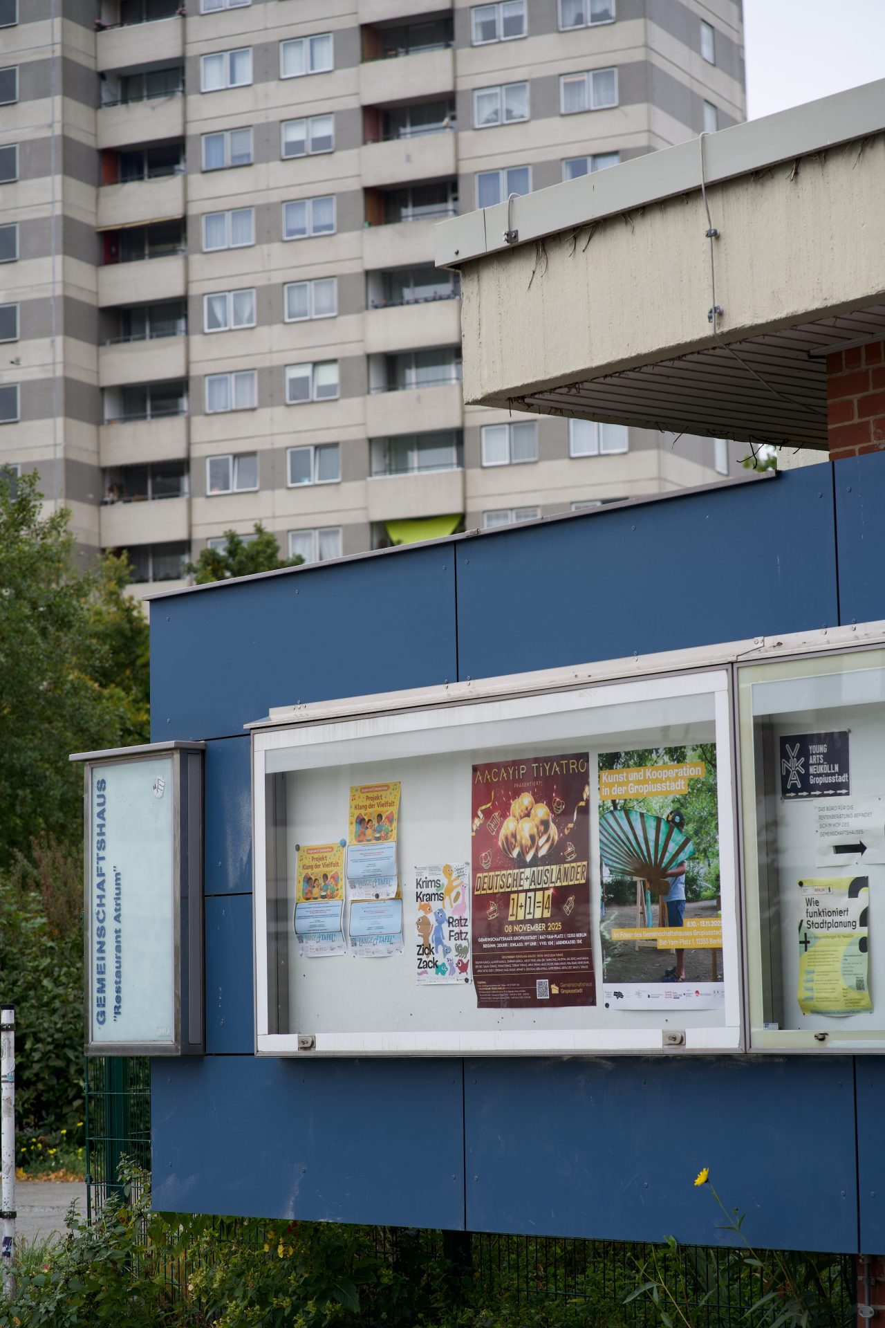 Plakatwand vor einem Gemeinschaftshaus mit verschiedenen Veranstaltungen, im Hintergrund ein mehrstöckiges Wohngebäude.