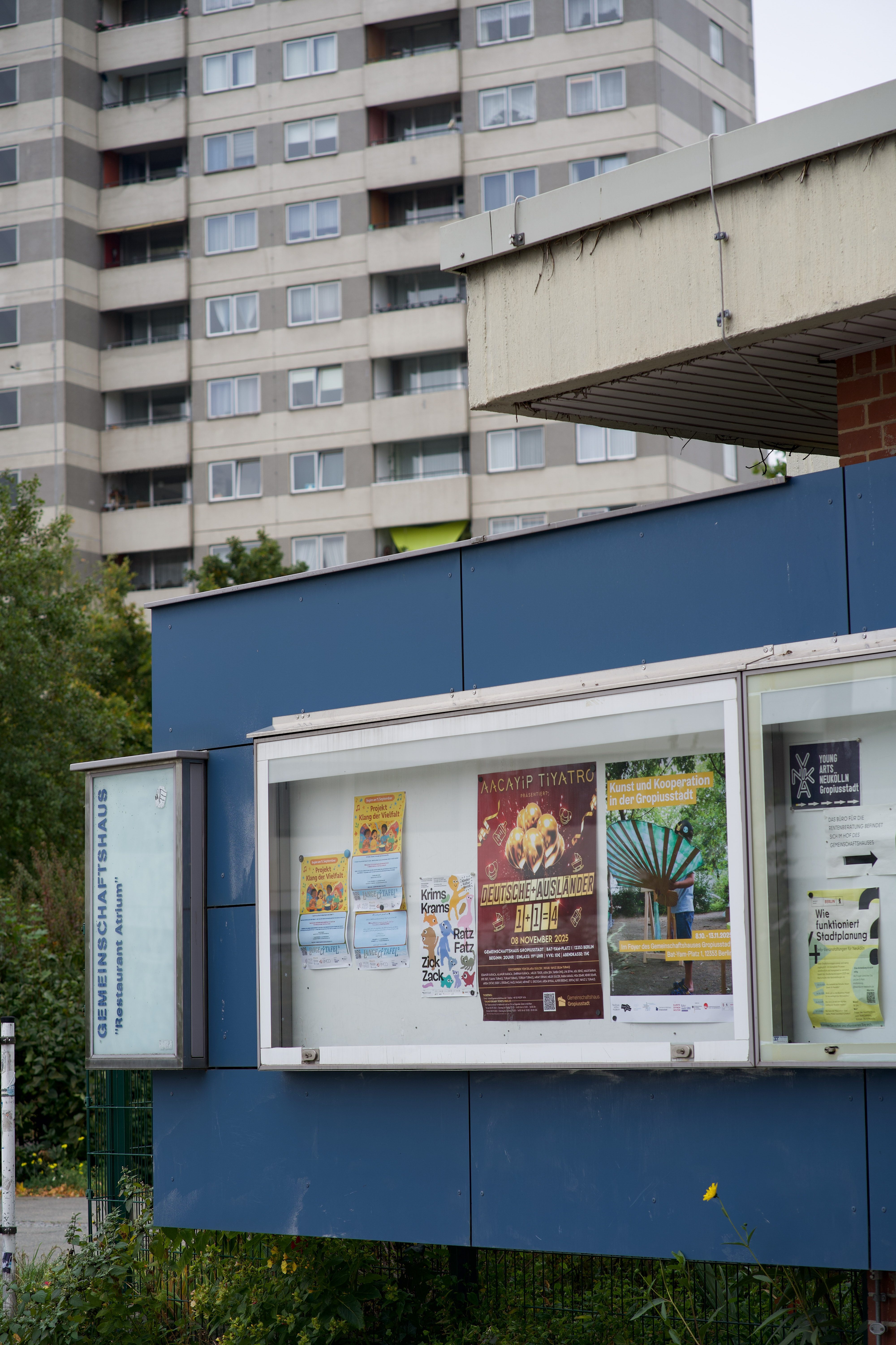 Plakatwand vor einem Gemeinschaftshaus mit verschiedenen Veranstaltungen, im Hintergrund ein mehrstöckiges Wohngebäude.