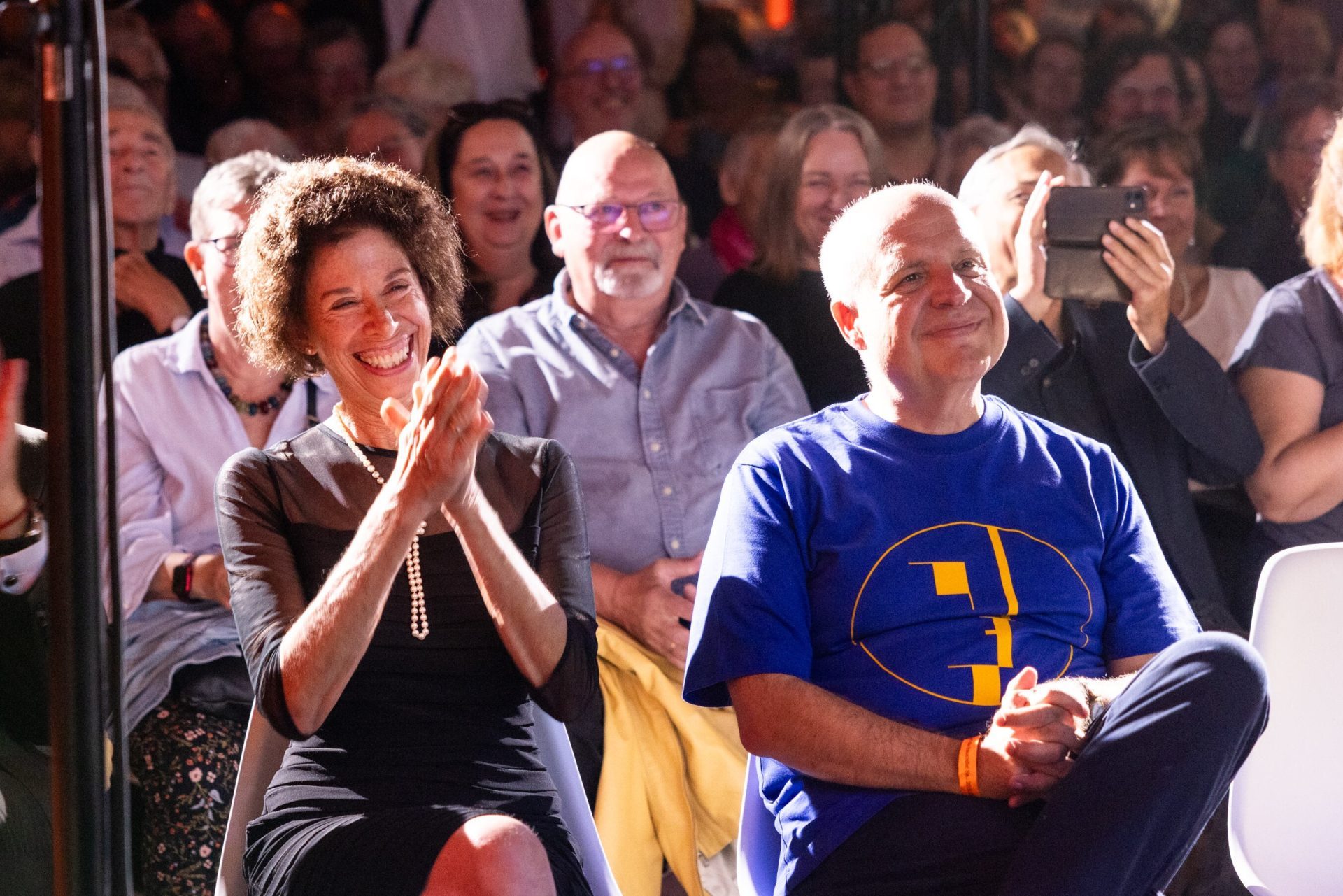 Satisfied audience members applaud at an event, smiling and attentive, in a well-filled hall.