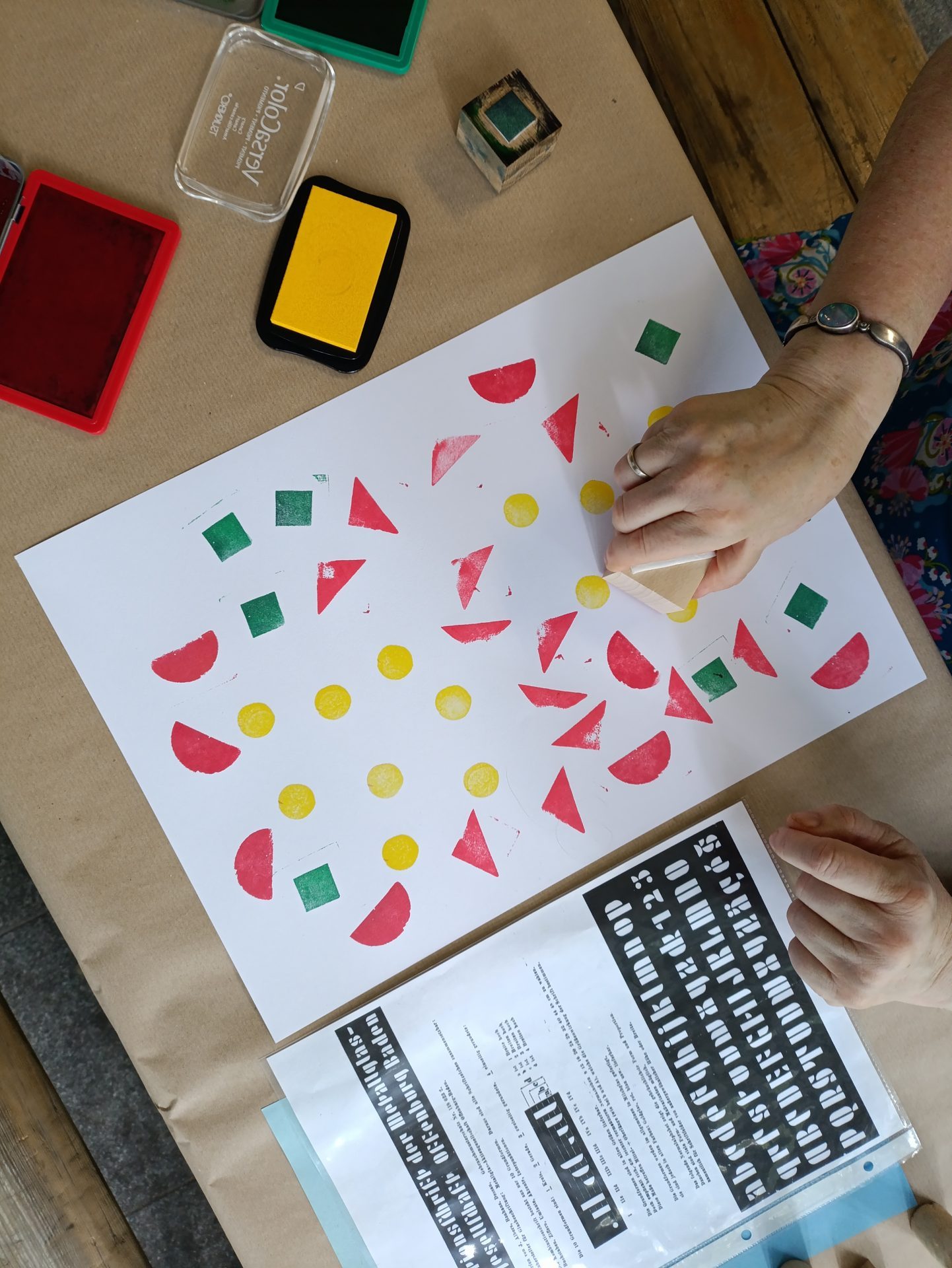 Auf dem Foto sind verschiedene Stempel zu sehen, außerdem eine Hand die auf einen Stempel auf ein bereits bunt bestempeltes weißes Blatt setzt.