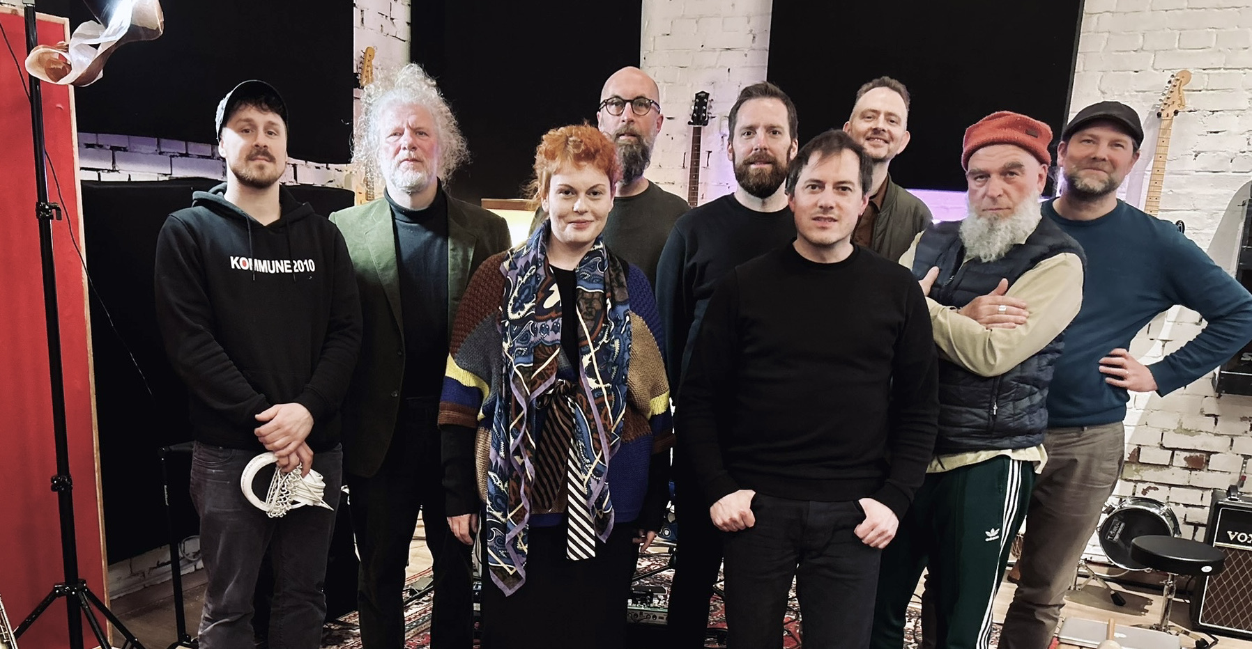 Eine Gruppe von neun Personen posiert in einem kreativen Studio mit Musikinstrumenten im Hintergrund.