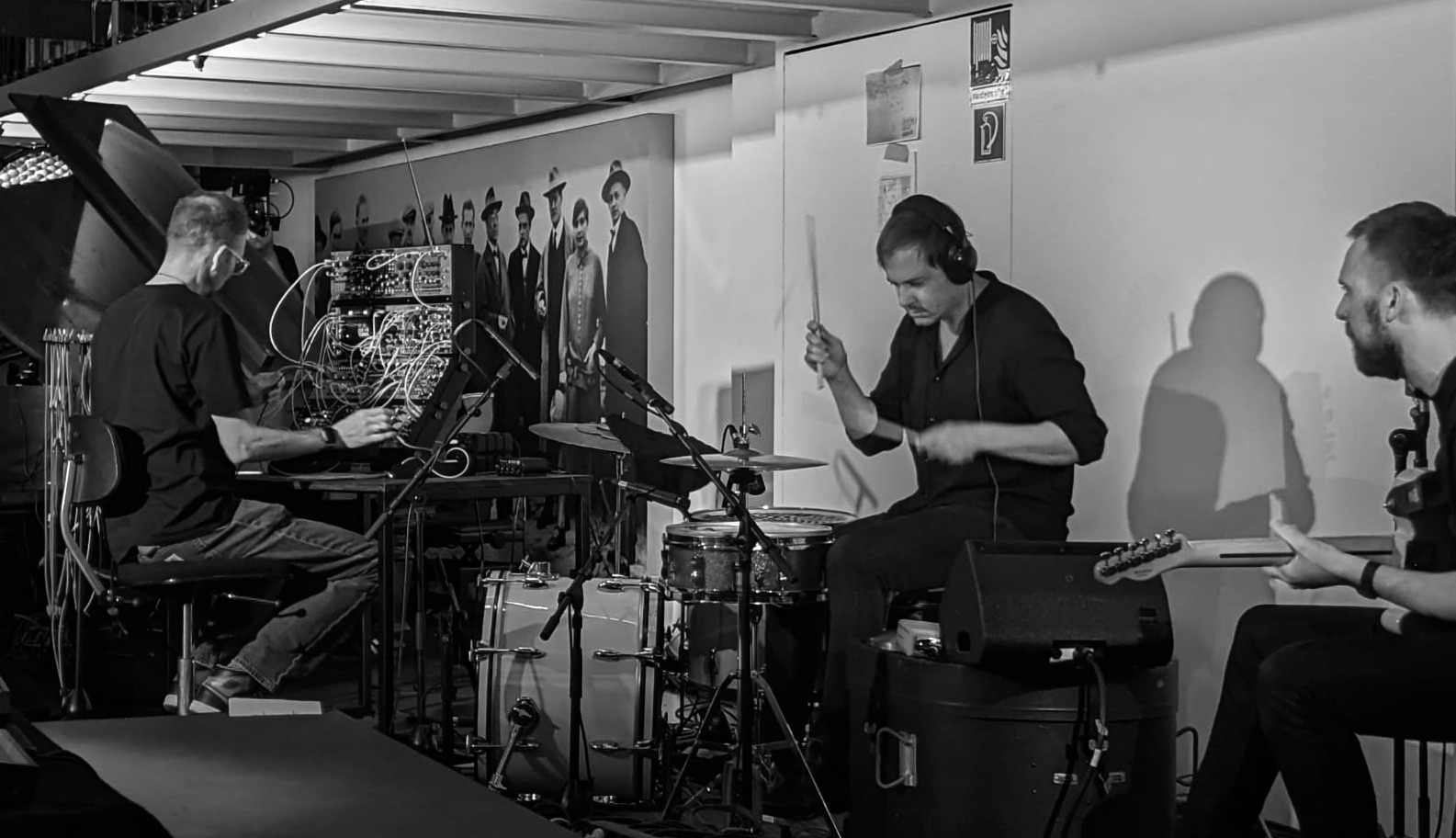 Drei Musiker spielen live: einer am Synthesizer, einer an den Drums, einer an der Gitarre, im Hintergrund schwarz-weiße Fotos.