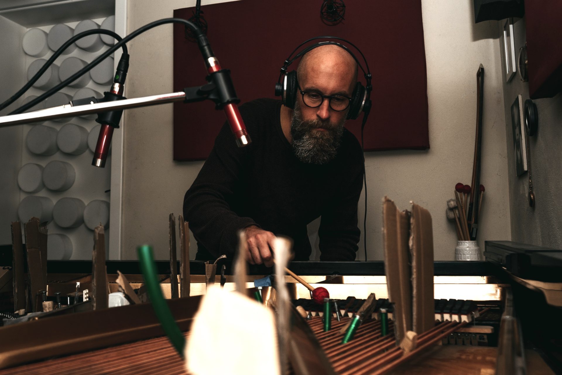 Mann mit Brille und Kopfhörern arbeitet an einem Tisch, im Hintergrund sind Musikinstrumente und Mikrofone sichtbar.