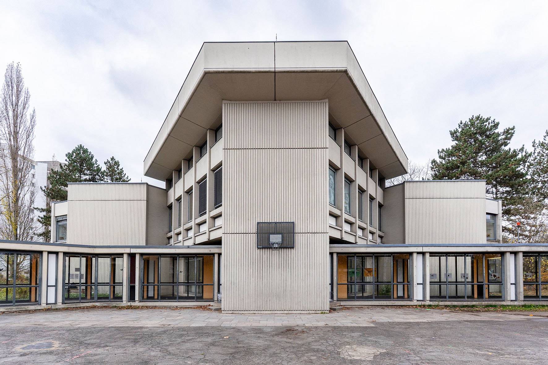 Moderne Architektur mit flachem Dach und großem Eingangsbereich, umgeben von Bäumen und einem grauen Himmel.