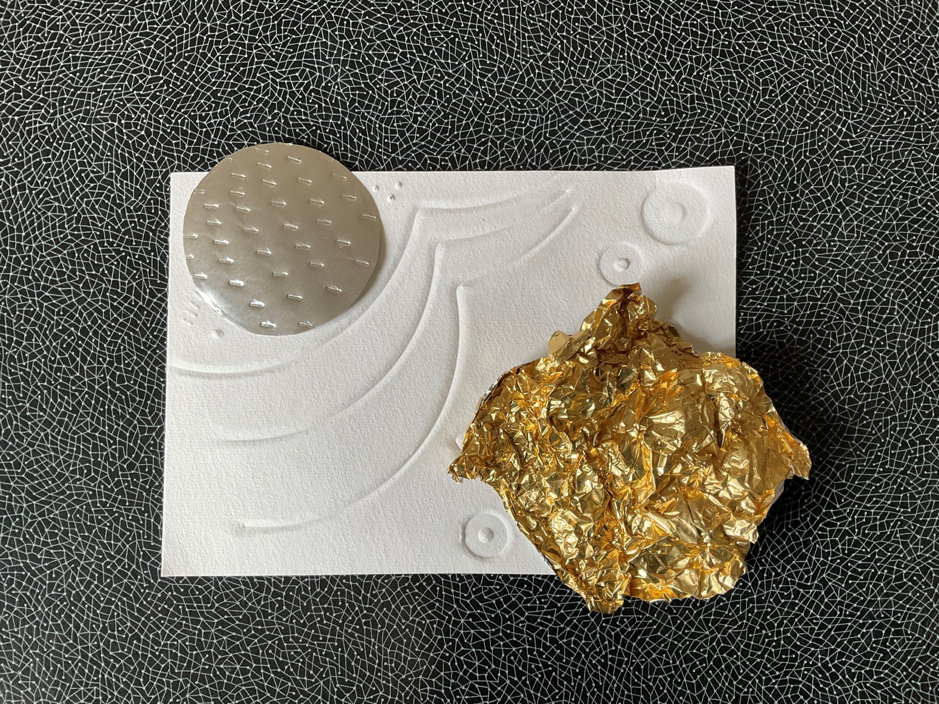Ein weißer rechteckiger Zettel liegt auf einem dunklen Untergrund. Auf der Oberfläche des Zettels drücken sich Kreise und Kurven ab. In der linken oberen Ecke des Zettels liegt ein Kreis aus silberner Folie. Auf der unteren rechten Ecke des Zettels liegt ein Stück zusammengeknüllte goldene Folie.
