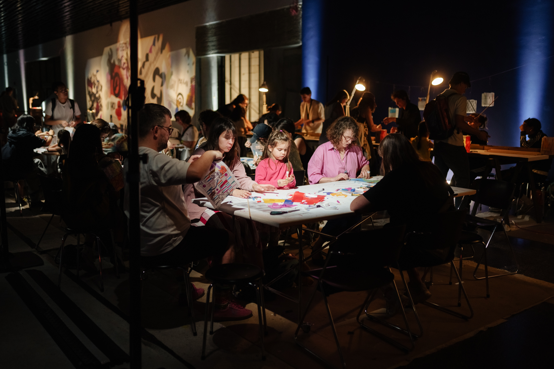 Gruppenausstellung mit Menschen, die kreativ an Tischen arbeiten, umgeben von bunter Kunst in einem gedämpften Raum.