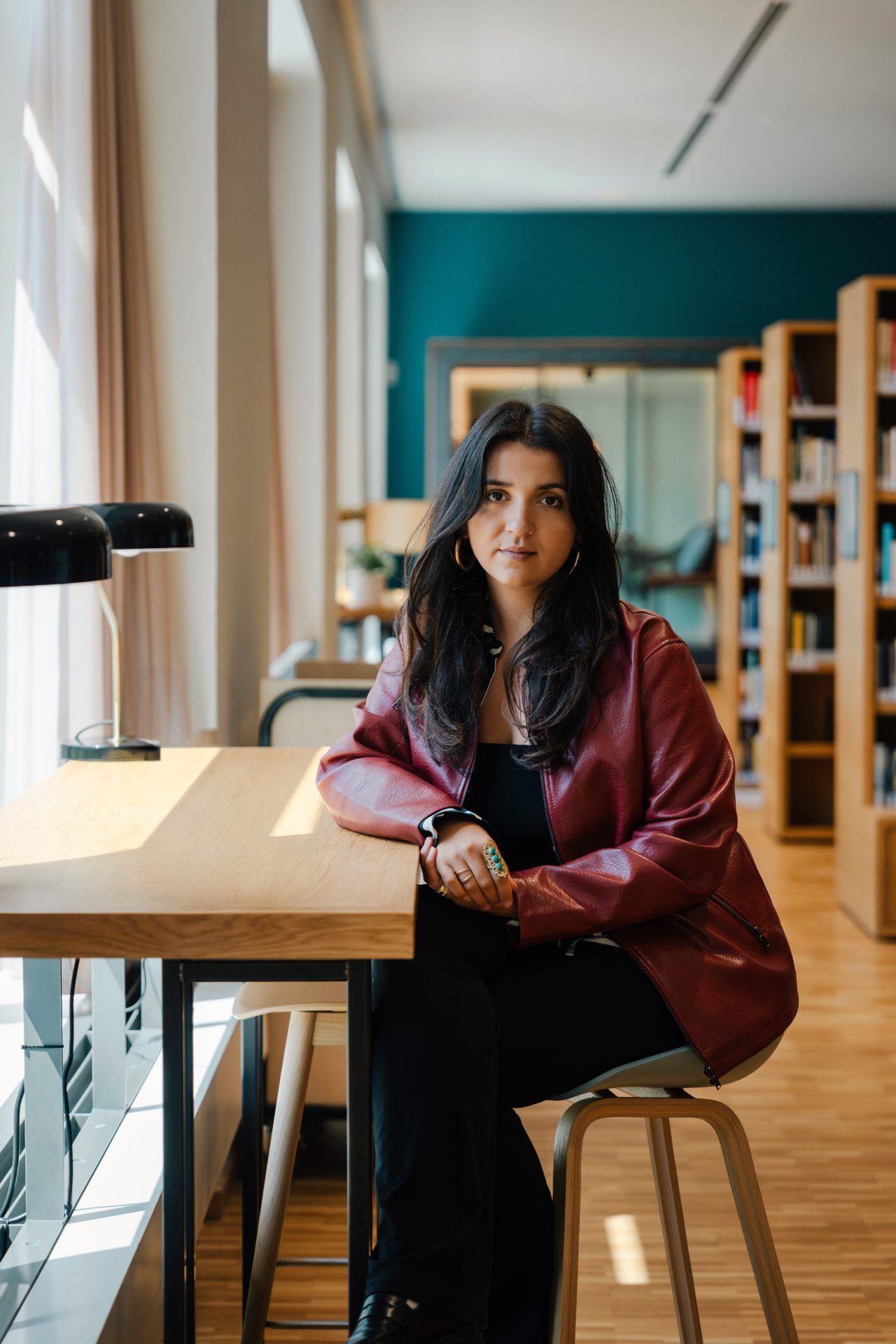 Eine Frau in roter Jacke sitzt an einem Tisch in einem hellen Raum mit Bücherregalen im Hintergrund.