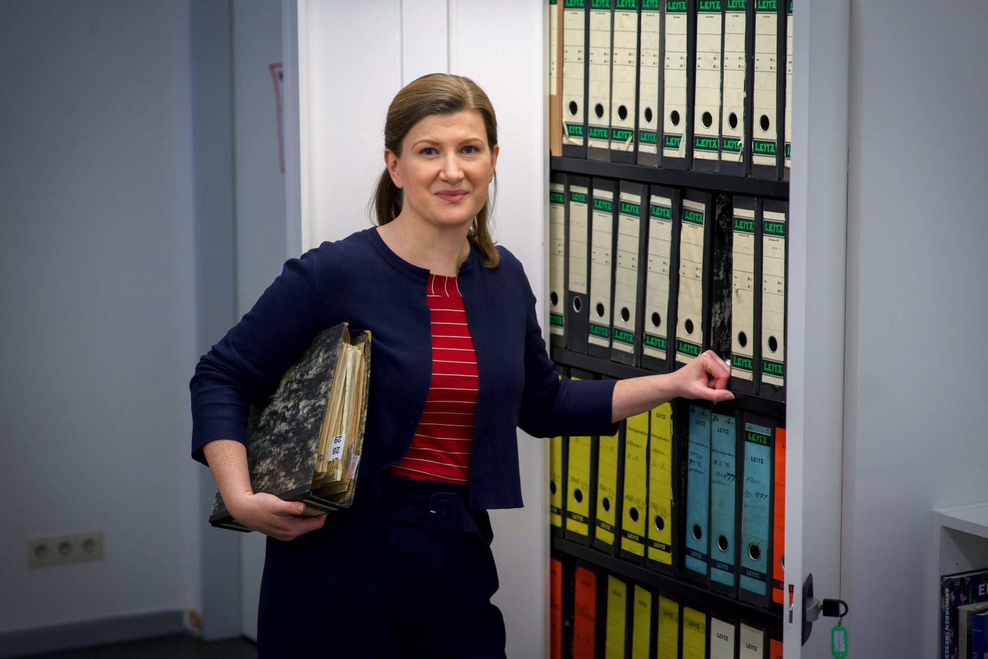 Frau mit Dokumenten steht vor einem Regal mit bunten Ordnern in einem Büro.