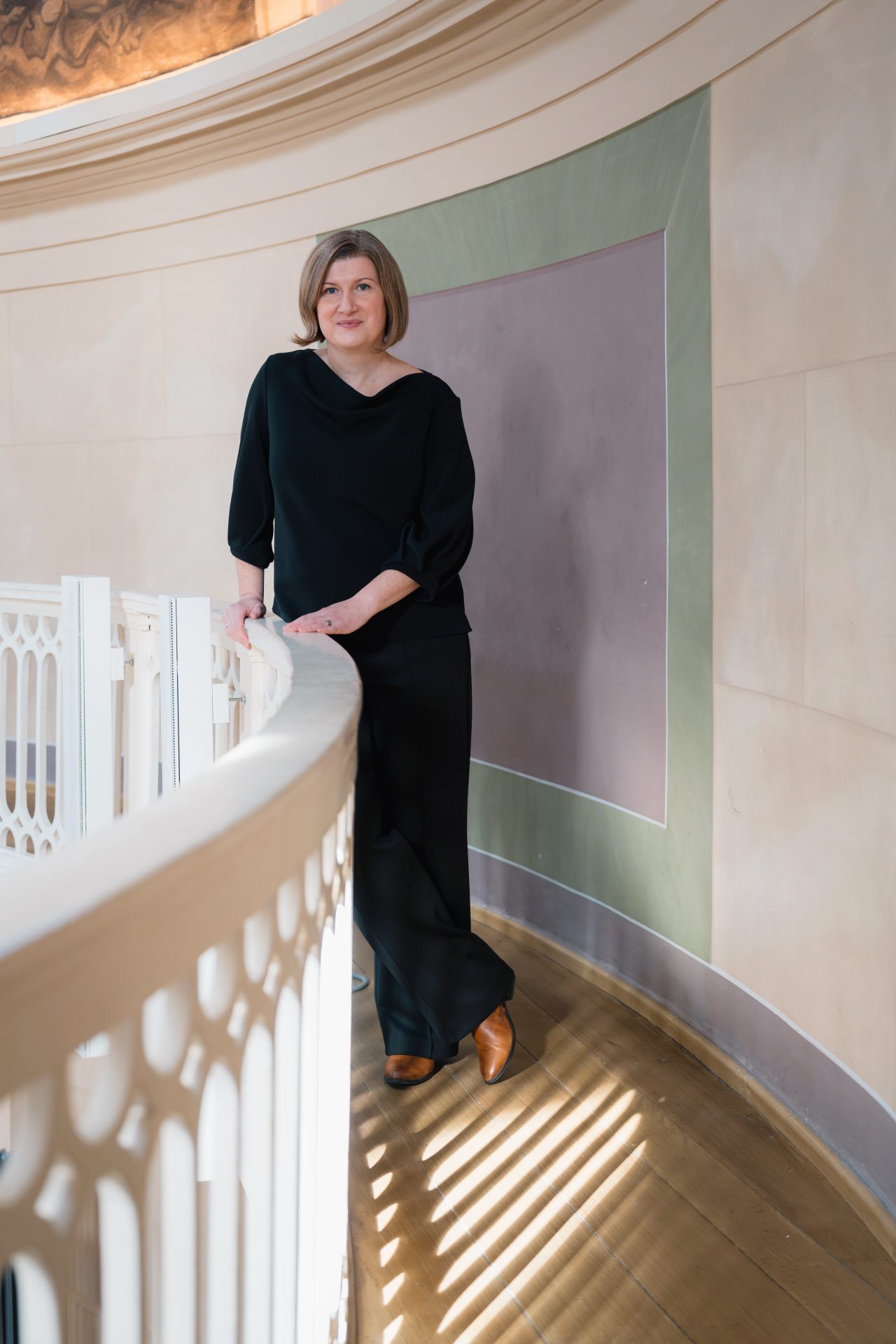 Frau in schwarzem Outfit lehnt an eleganter Balustrade in einem hellen, minimalistischen Raum mit Holzfußboden.